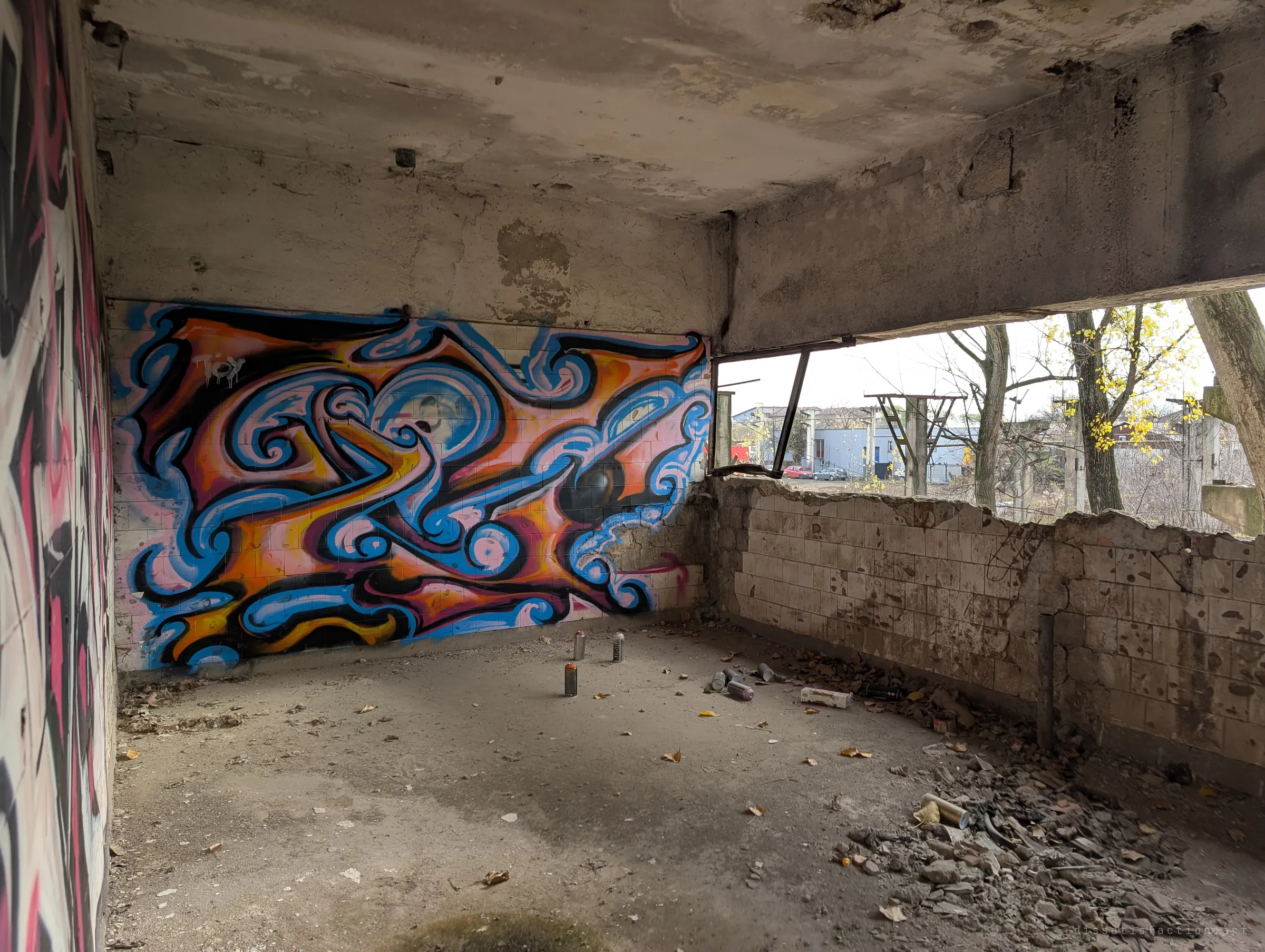 A small concrete room with a vivid blue and orange graffiti piece, spray cans still standing on the floor. The broken window opens onto trees and neighbouring buildings.