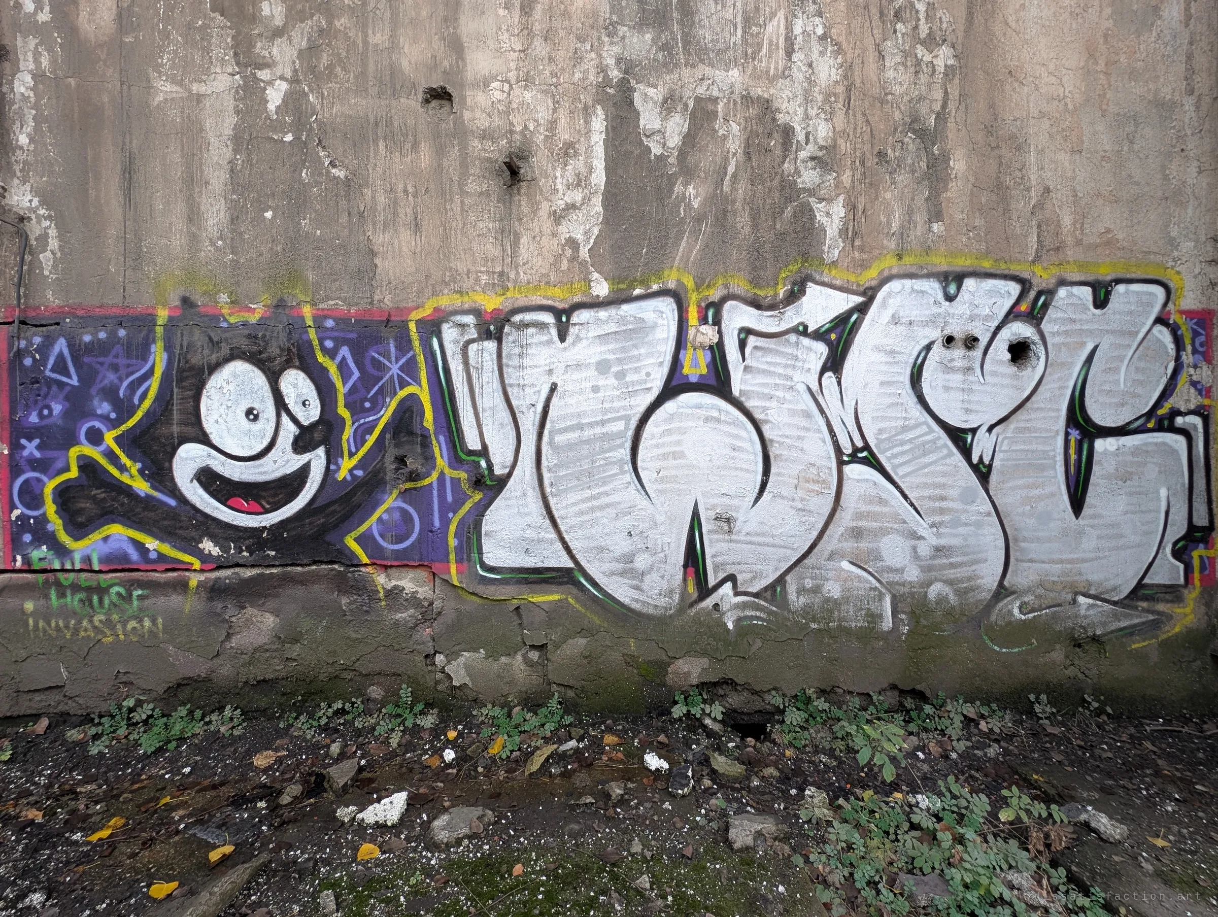 A cartoon cat and a large silver graffiti piece run along the base of a weathered concrete wall, with weeds and rubble in front. Street art has claimed the factory's outer walls.