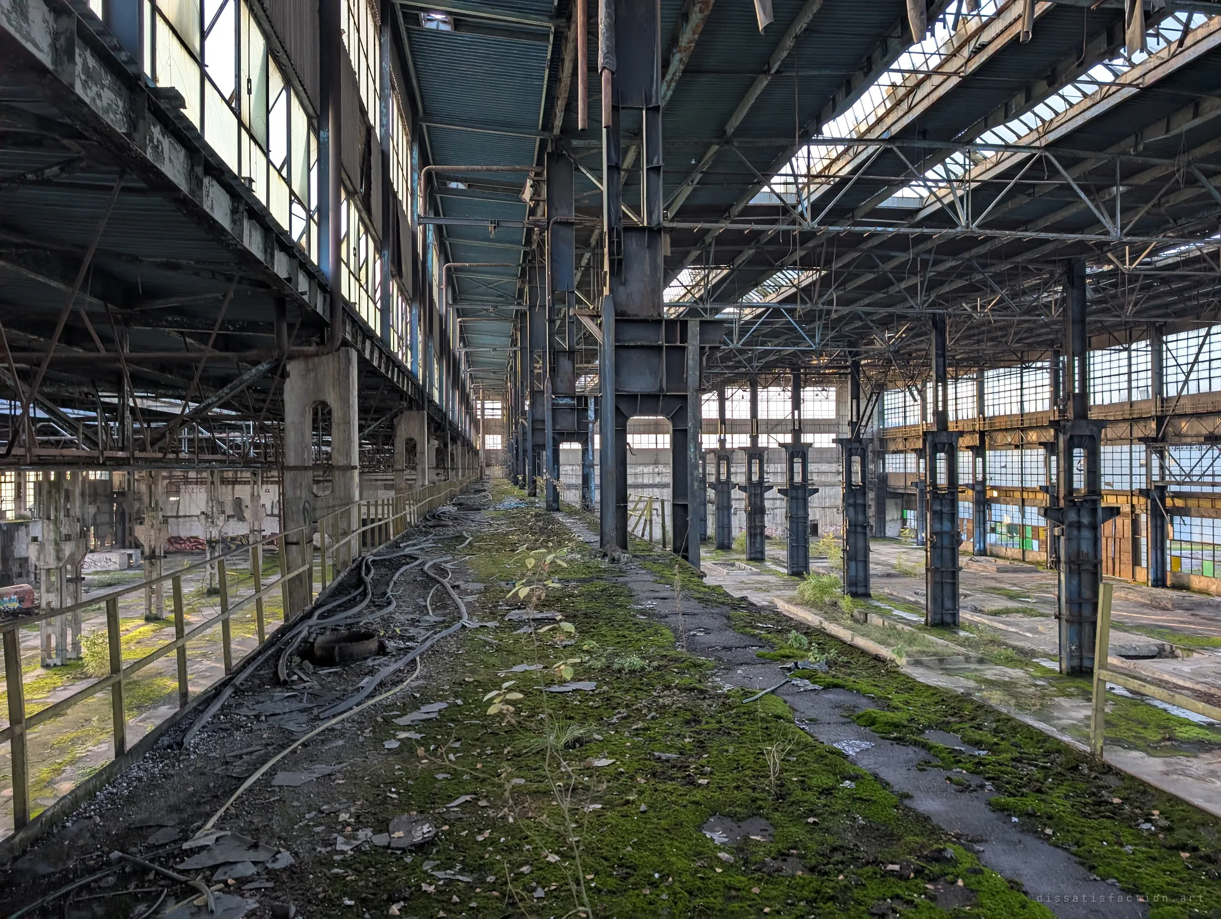 From the elevated gantry, a mossy walkway strewn with old cables stretches into the distance beside a row of massive steel crane columns. Daylight pours through the torn roof onto the empty factory floor.