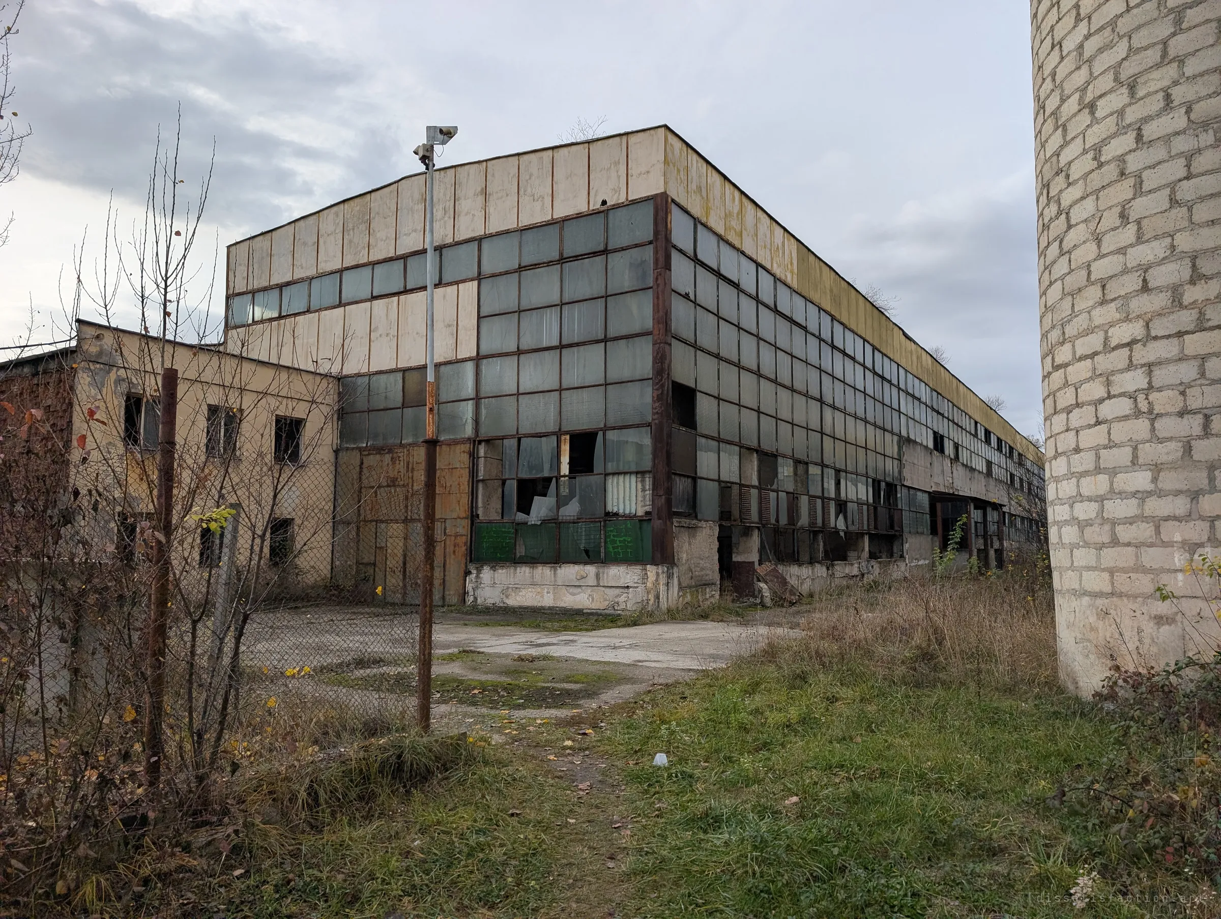 A long hall of cracked glass panes stretches away under a grey sky, fronted by overgrown grass and a rusty fence, with a brick chimney at the edge. The outside view shows the scale of the abandoned works.