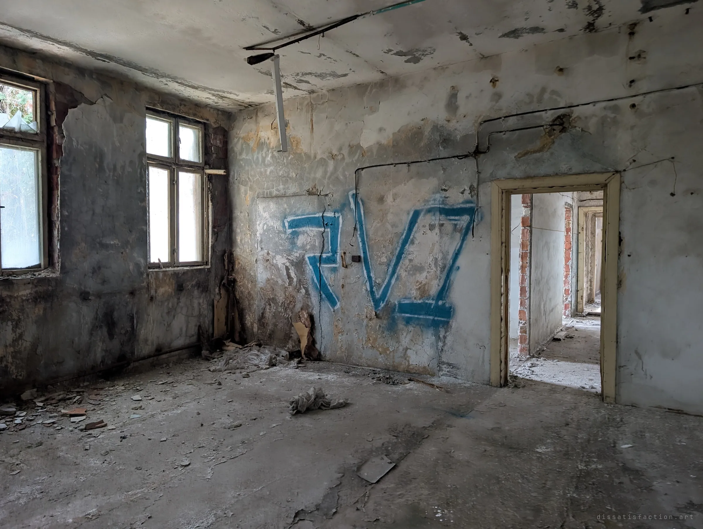 An empty office room with peeling grey walls and a blue graffiti tag, its doorway opening onto a line of stripped rooms. The dangling light fitting and debris show years of neglect.