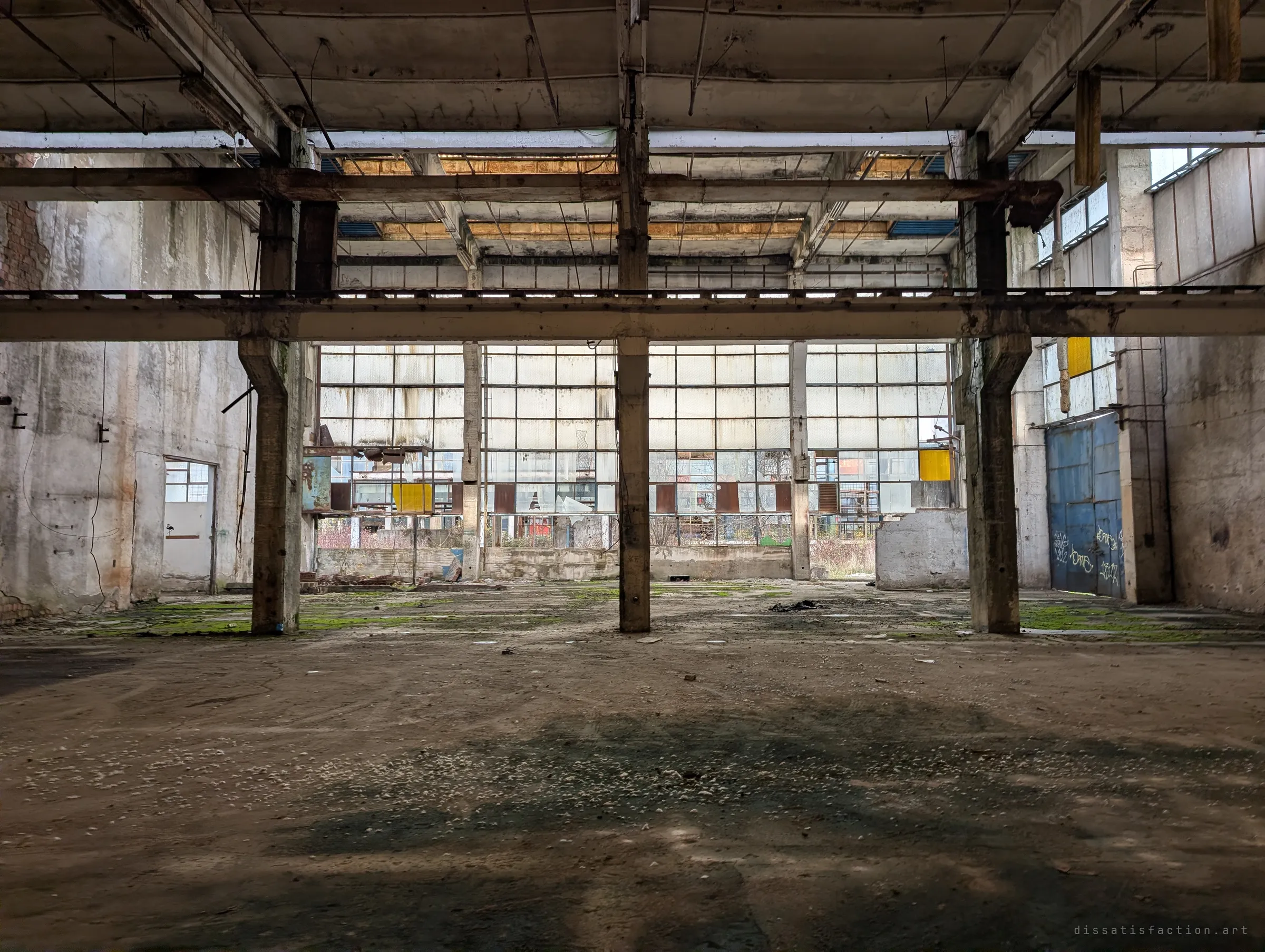Concrete columns and a crane rail frame a wall of dusty glass in a vast, empty workshop floor patched with moss. The sheer volume of the space dwarfs everything in it.