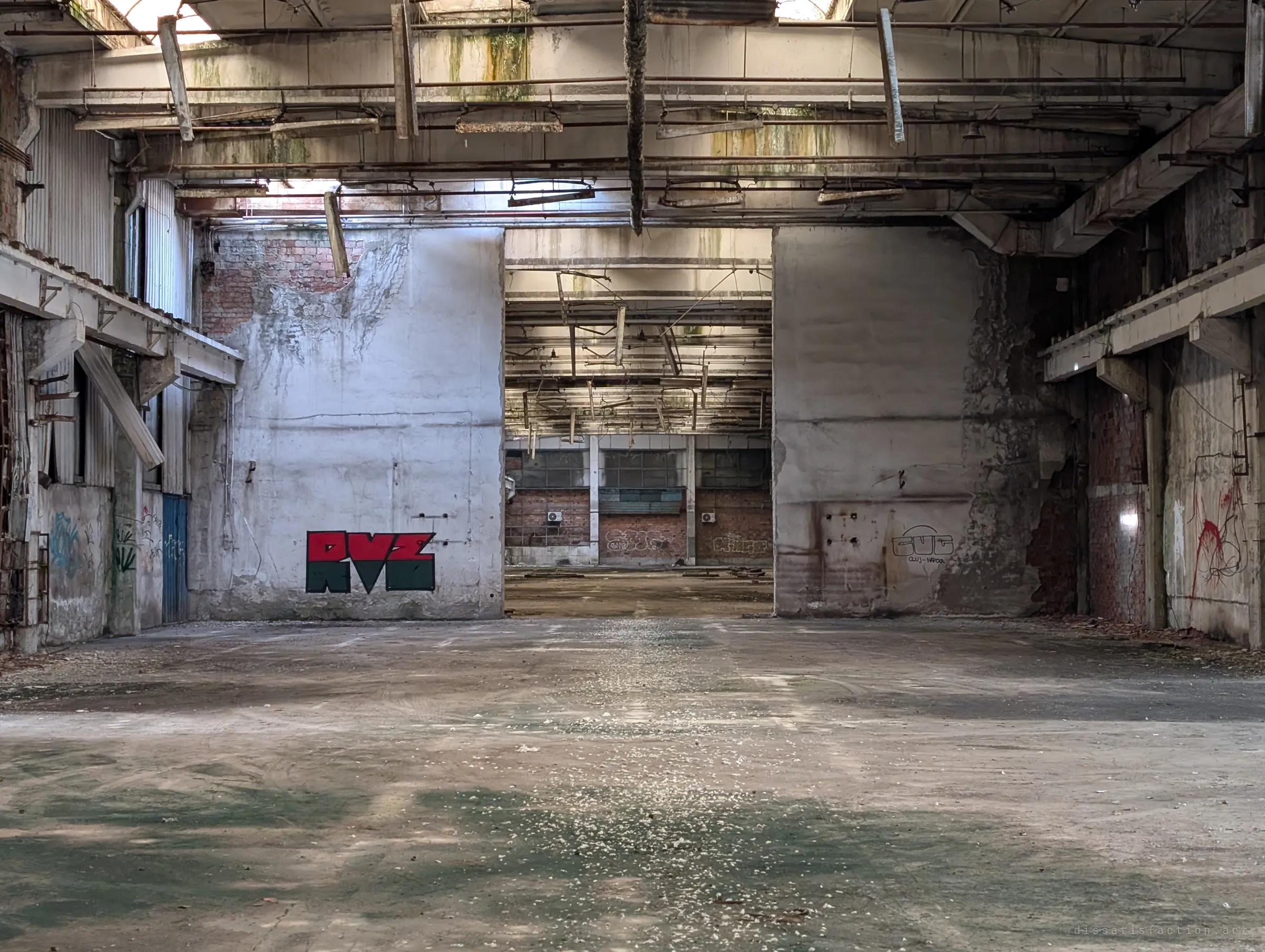A red and black graffiti piece sits on a white wall beside a wide opening into the next hall, where rows of hanging lights fade into the distance. The halls chain together through the whole building.