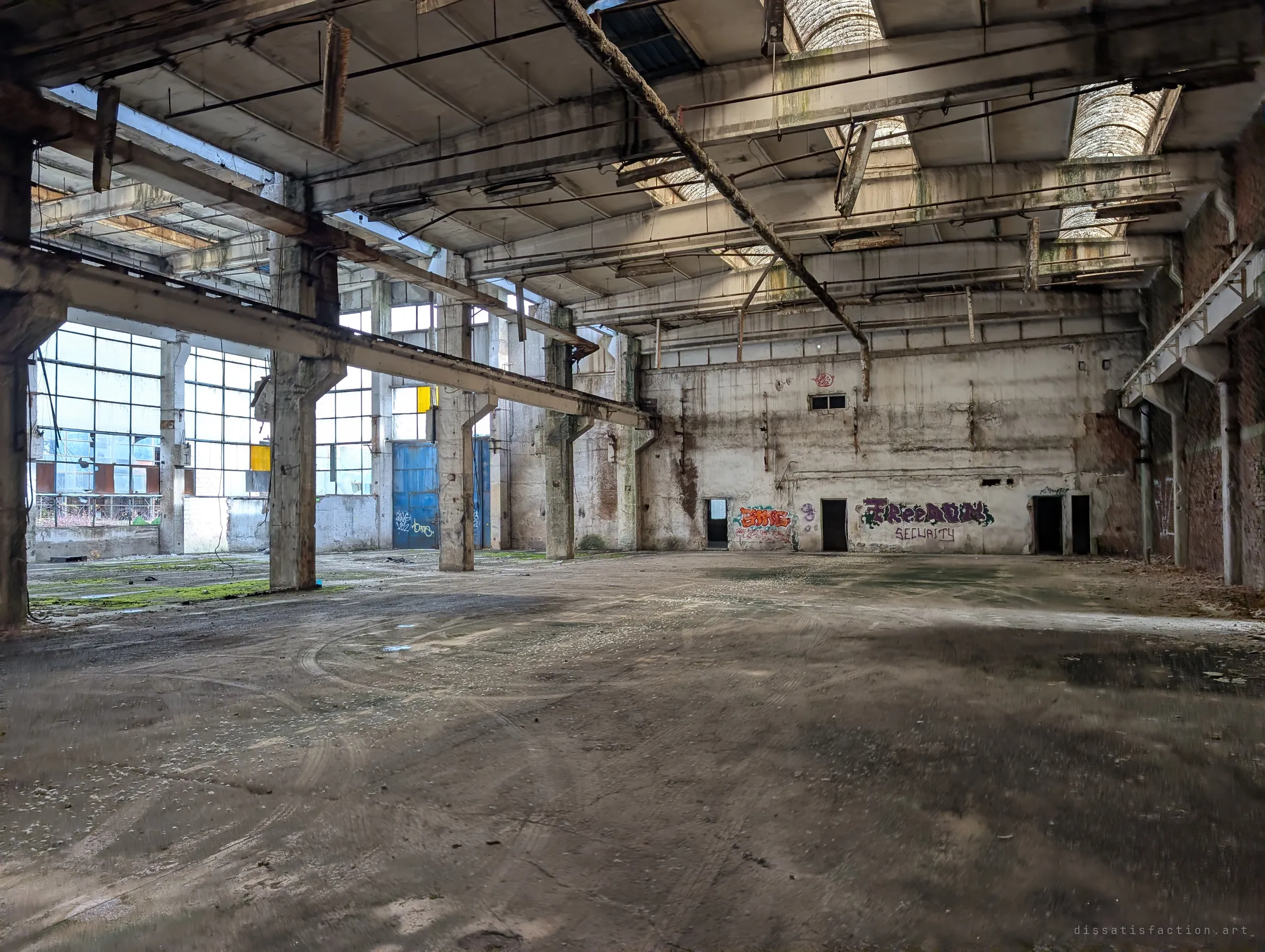 A cavernous workshop with fallen ceiling lights, tyre marks across the dusty floor and graffiti along the far wall. Blue doors and glazing line the left side.