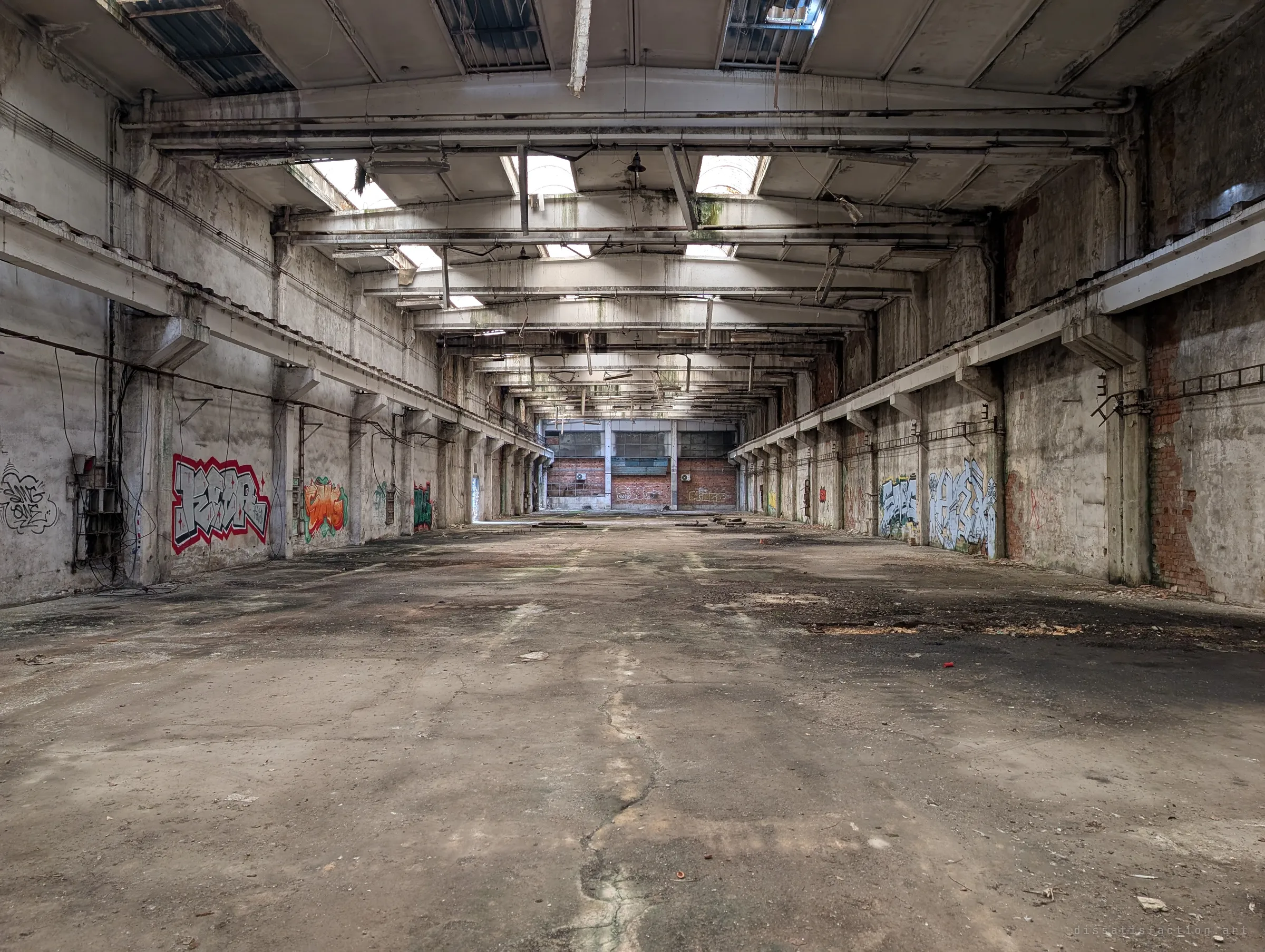 A long production nave stretches to a brick end wall, with skylights overhead and graffiti pieces along both sides. The symmetry pulls the eye down the length of the hall.