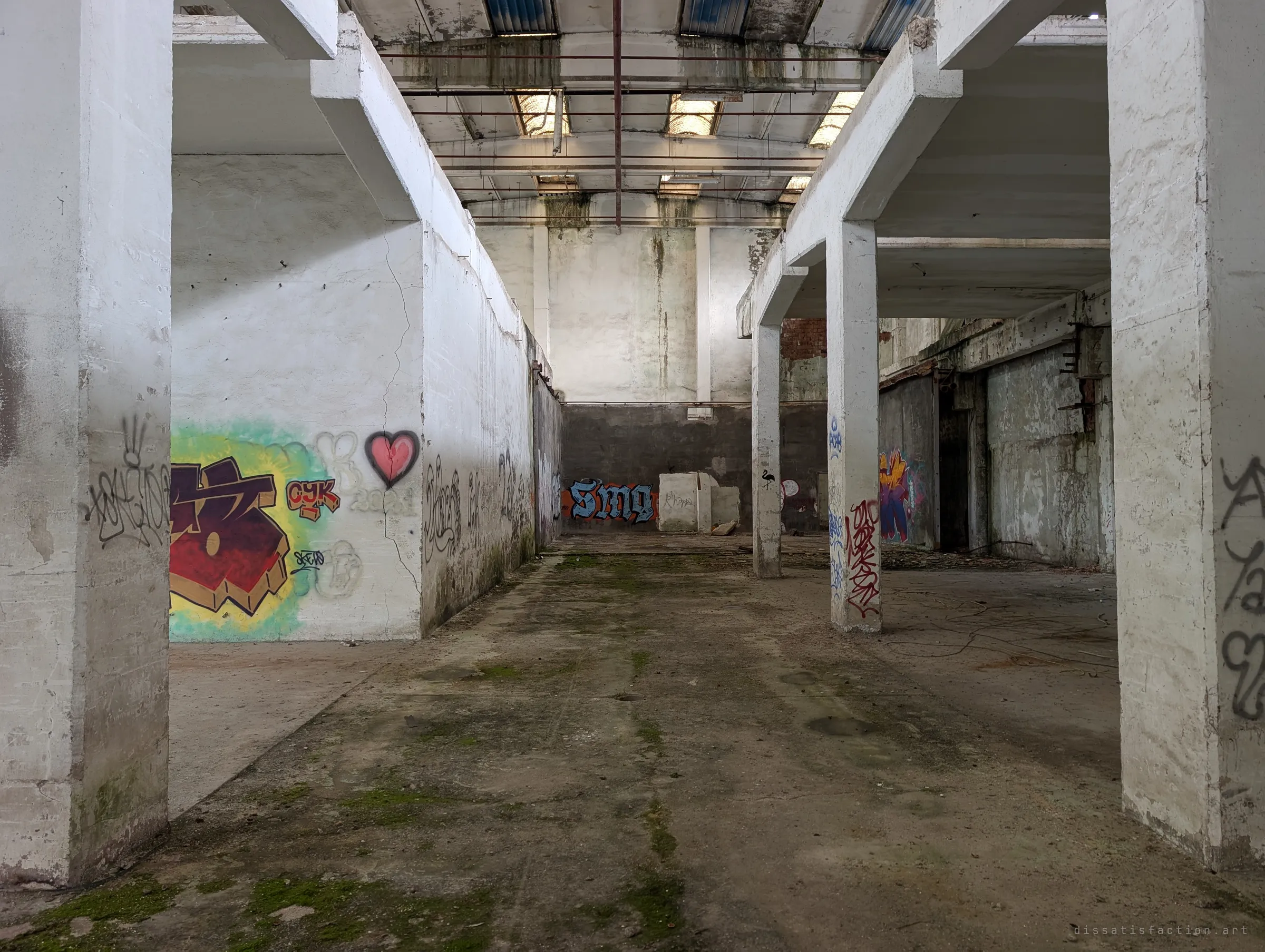 White pillars frame a bay covered in colourful graffiti, including a pink heart, beneath a roof of dirty skylights. Taggers have turned the empty hall into a gallery.