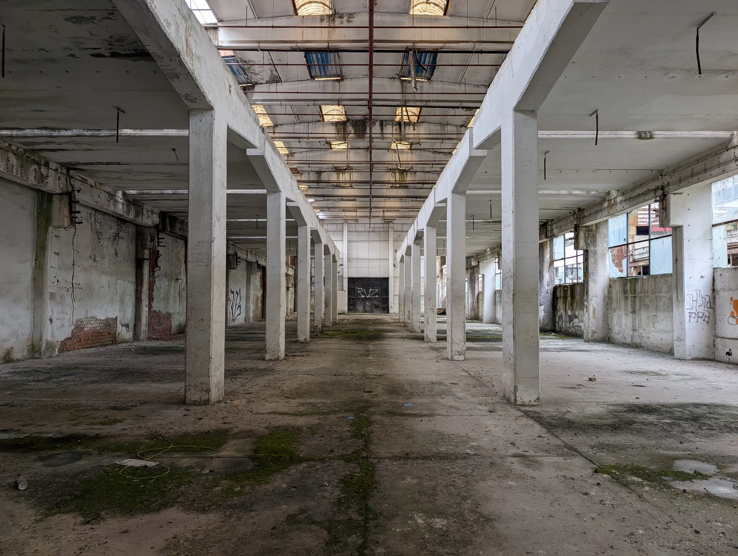 Two rows of white columns march down a moss-streaked hall toward a dark steel door marked with a tag. Soft skylight light fills the symmetrical space.
