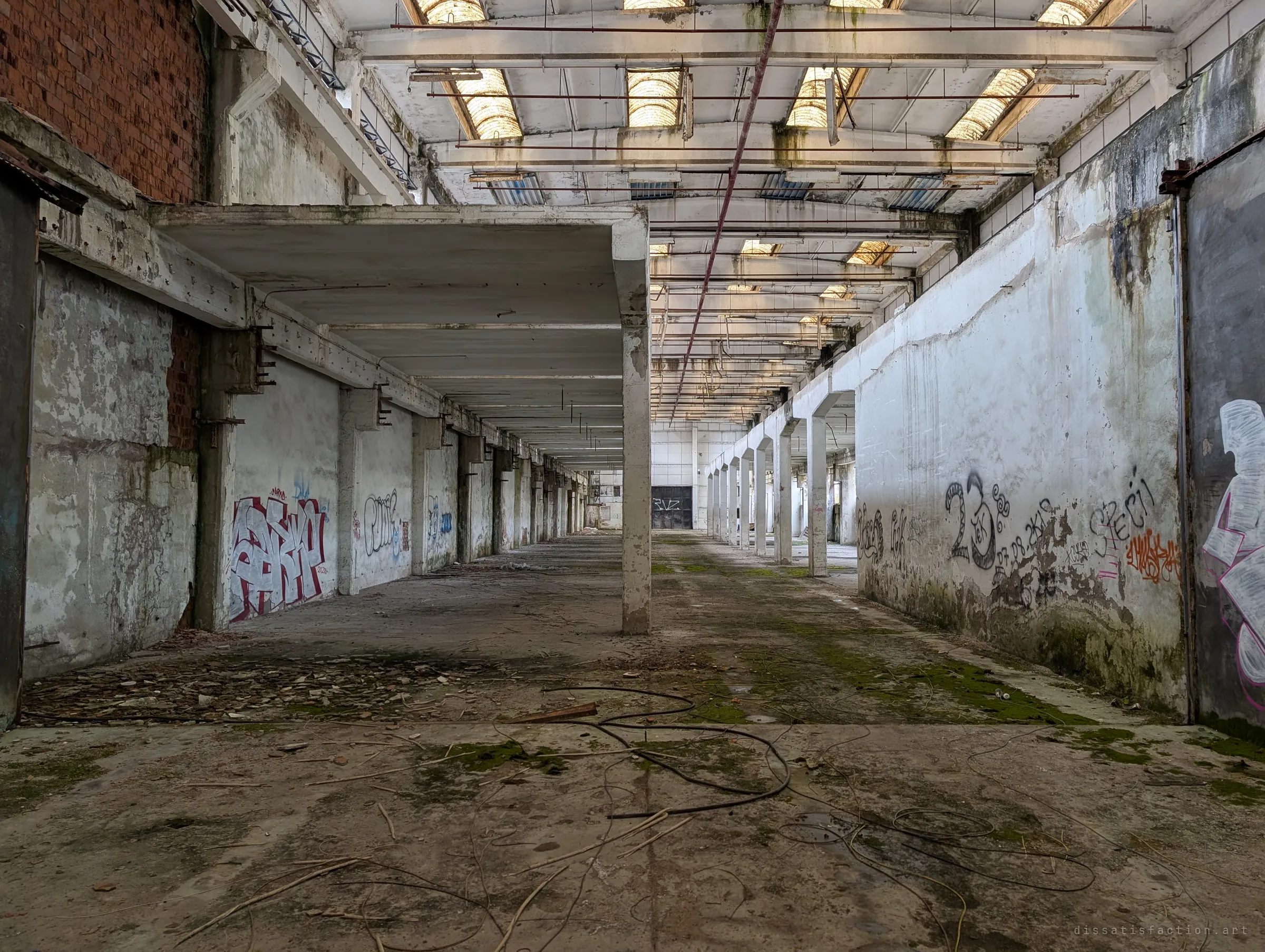 A long production hall stretches into the distance under rows of skylights, its walls tagged with graffiti and its floor strewn with old cable and moss. The repeating columns show the sheer scale of the abandoned plant.