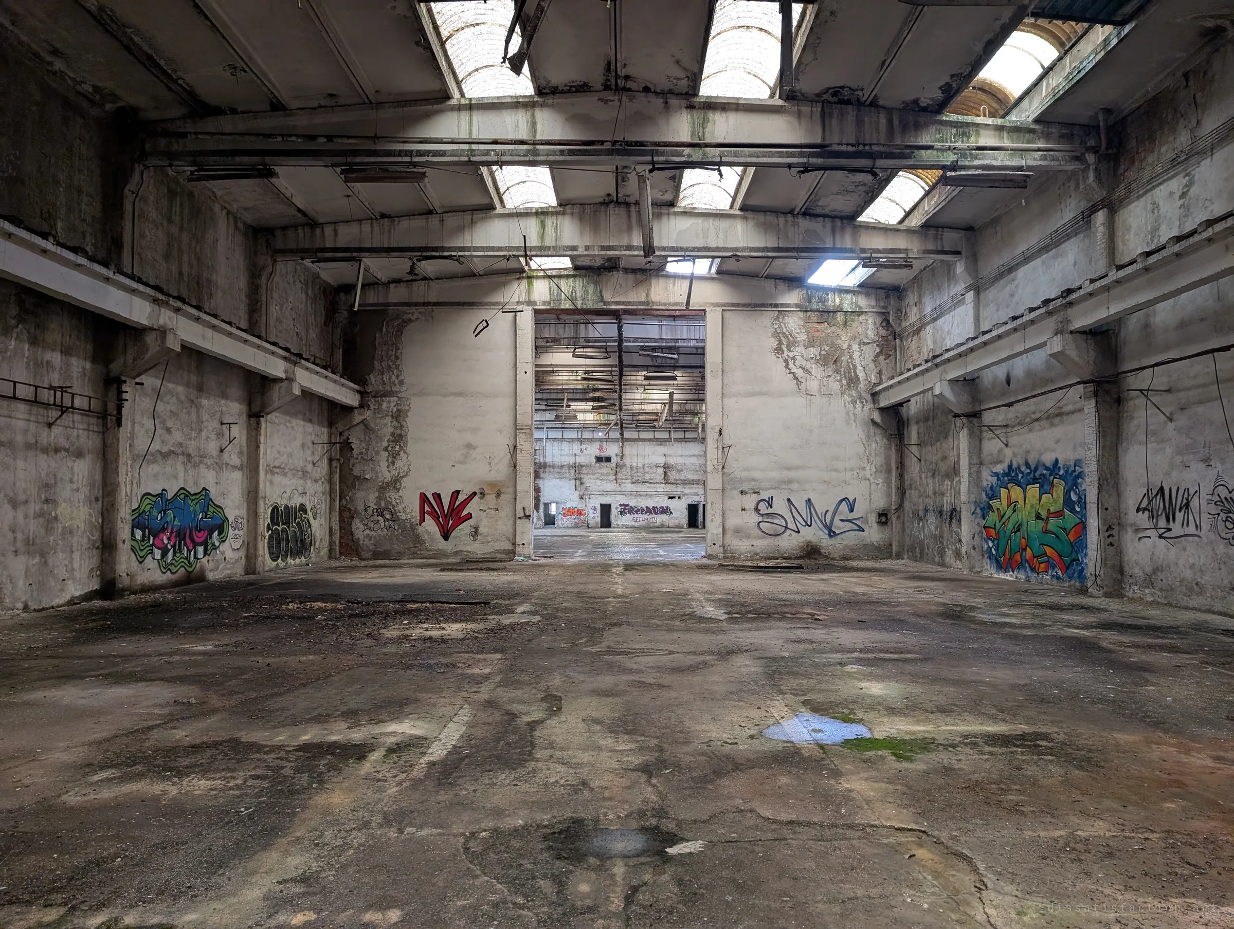 An empty bay with graffiti pieces on every wall opens through a tall doorway into another hall beyond. Daylight falls through the broken roof lights onto the cracked, puddled concrete.