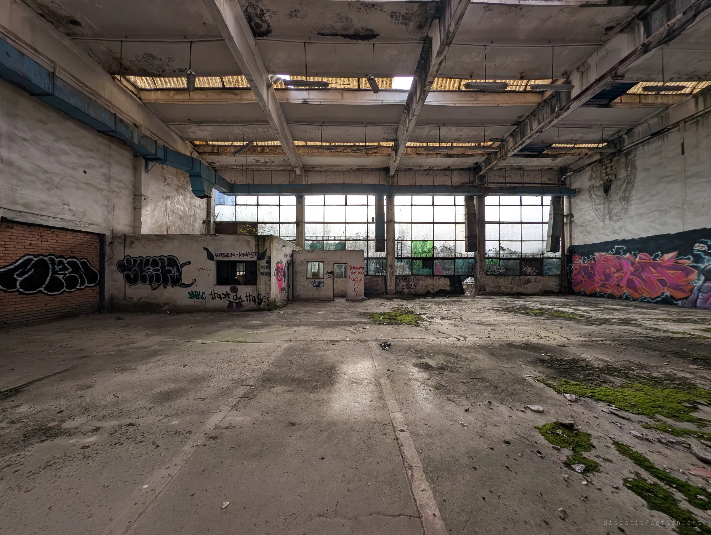 A wide workshop hall with a small office box built inside it, a glazed back wall and graffiti along both sides. Moss is taking over the bare floor under the sagging roof panels.