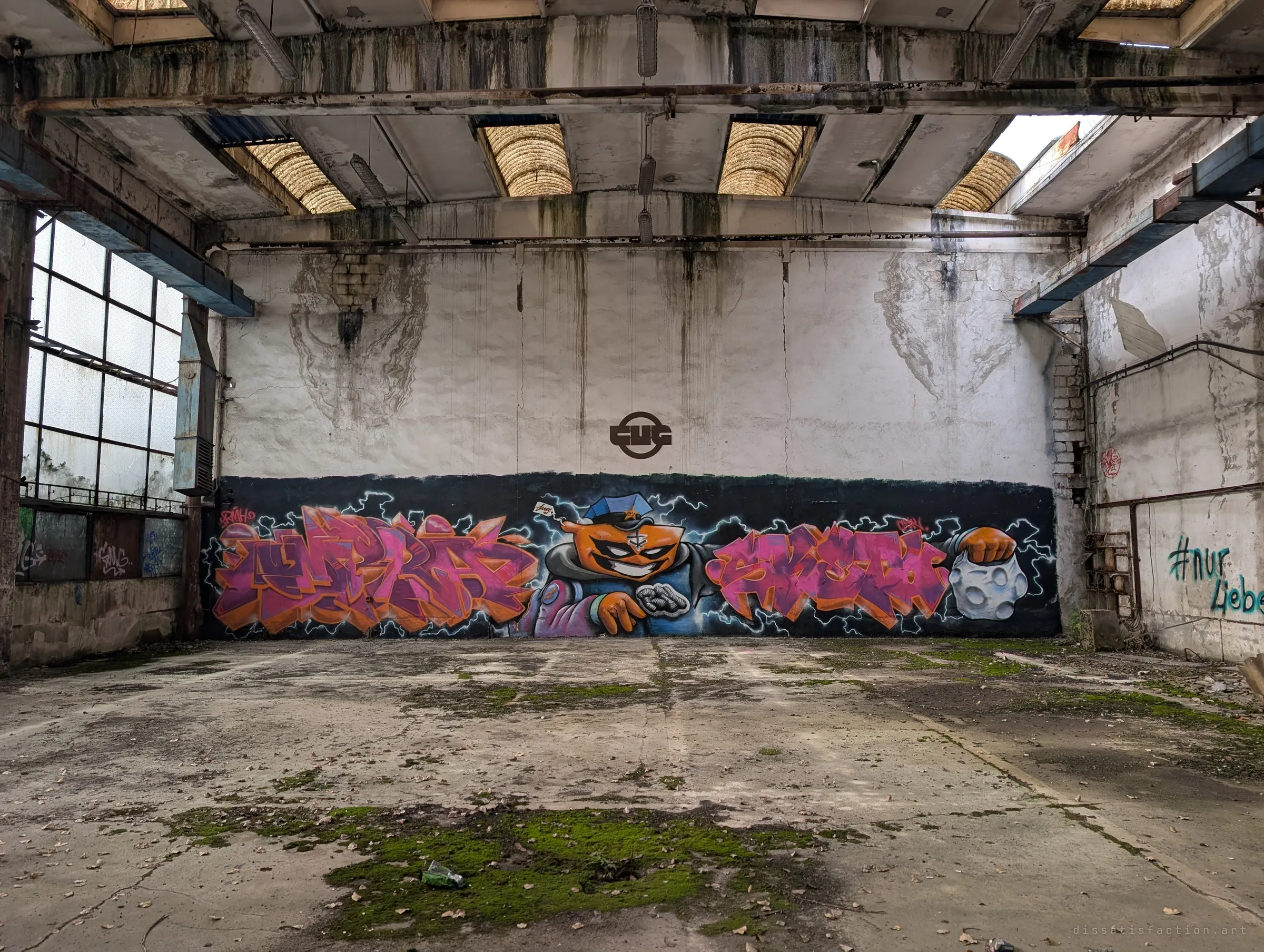 A large colourful graffiti mural with a grinning cartoon character spans the back wall of an empty hall, beneath the painted emblem of the former factory. Skylights and old ducting frame the scene above a mossy floor.