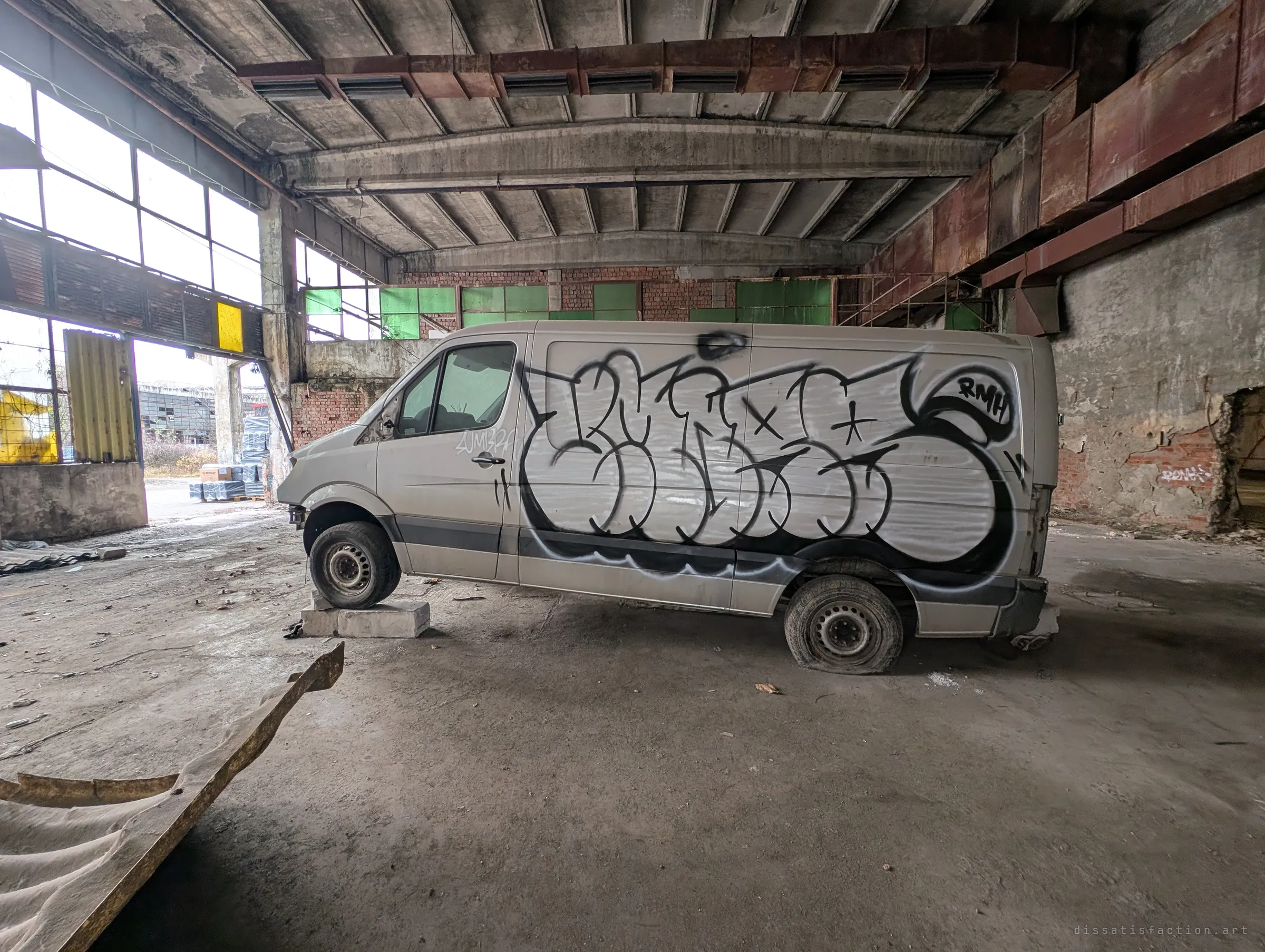 An abandoned van covered in a large black-and-white graffiti piece sits propped on concrete blocks inside a hall. Its flat tyres and the open wall behind it show how long it has been left here.
