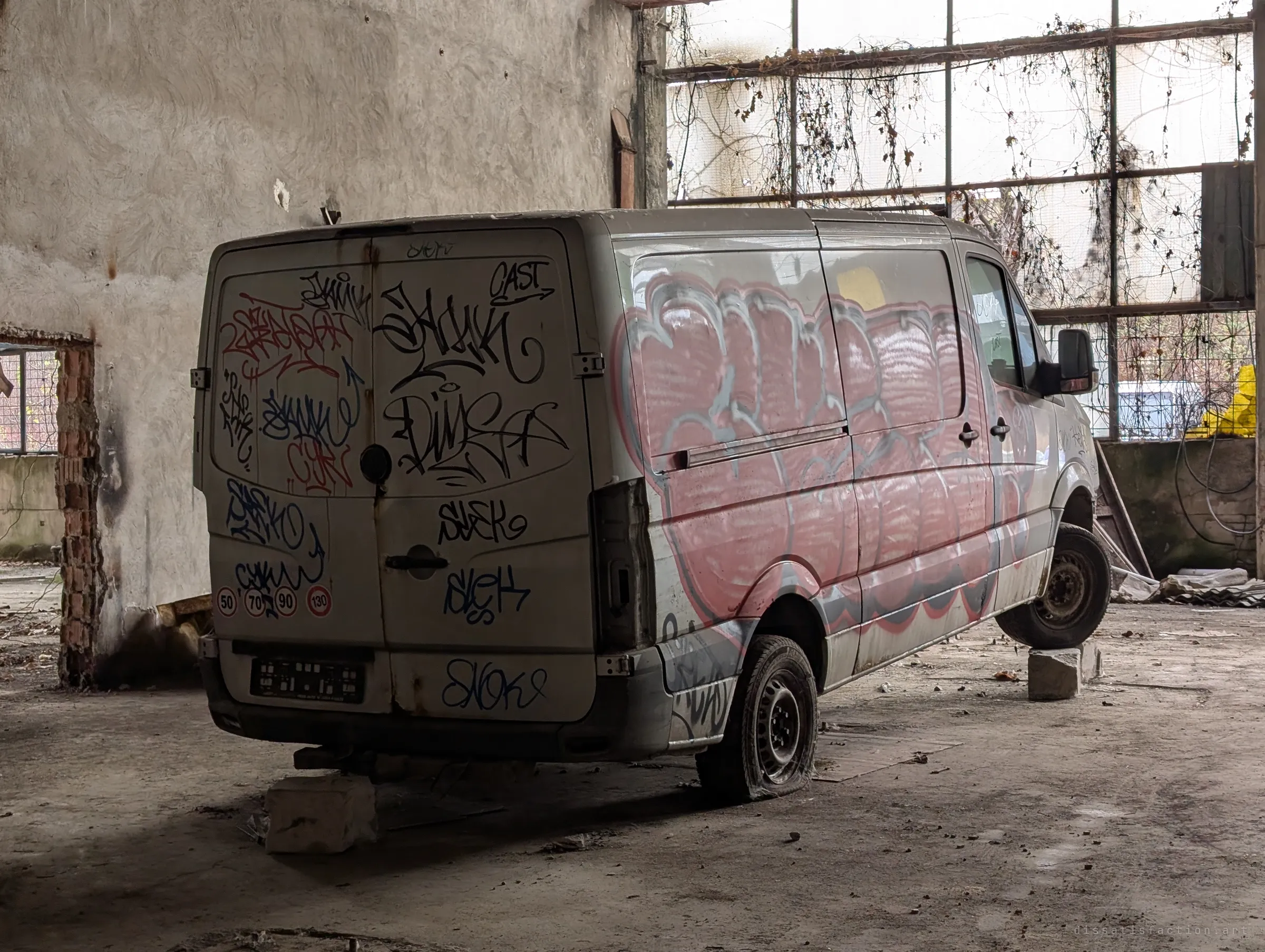 The rear of the same abandoned van, its doors covered in tags and its side in a faded pink throw-up, resting on blocks with flat tyres. Vines hang over the tall grid windows behind it.
