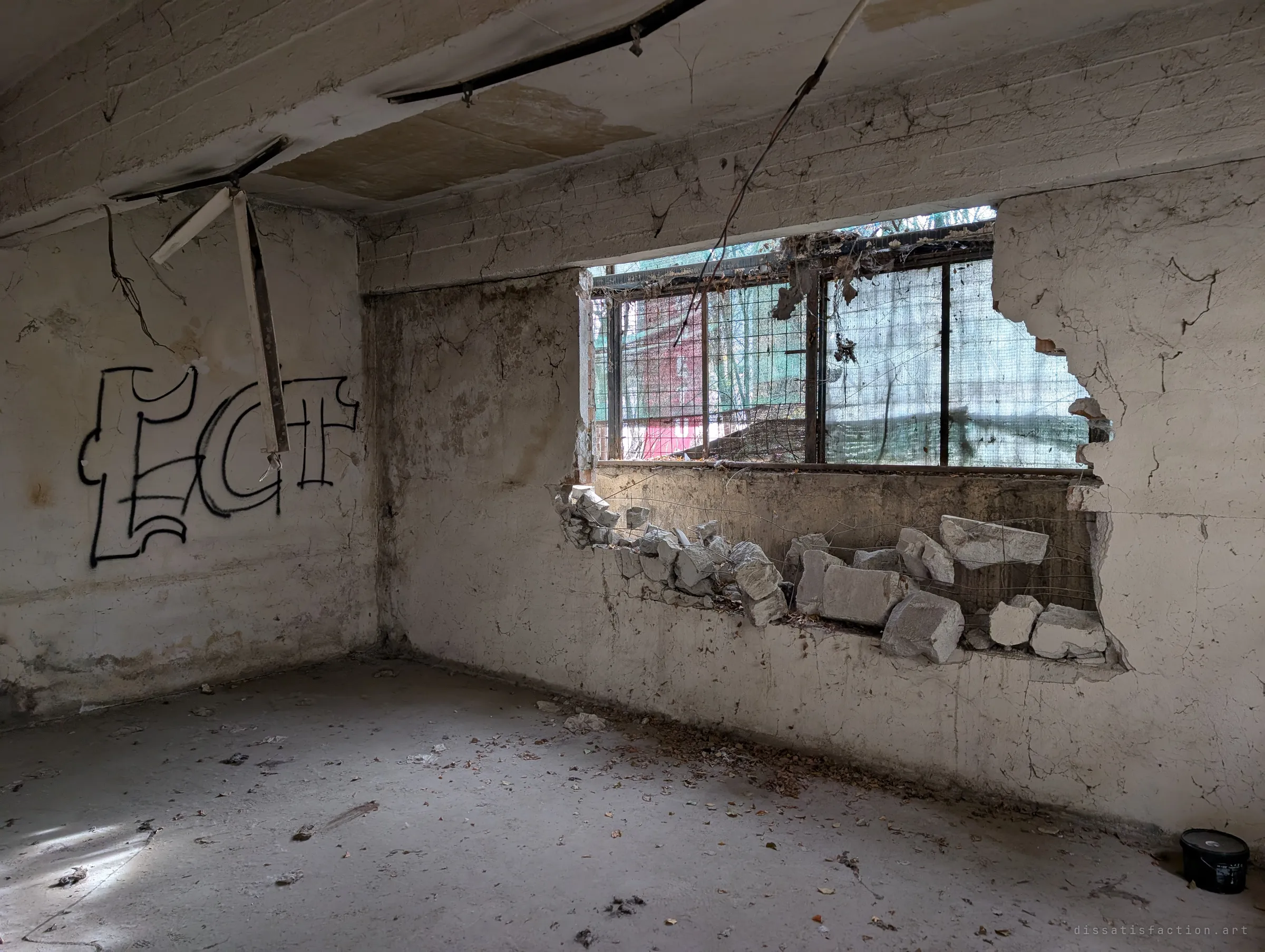 A small side room with a window hole broken out of its wall, rubble piled on the sill and a single graffiti tag on the wall. Light filters in through a wire-mesh window beyond.