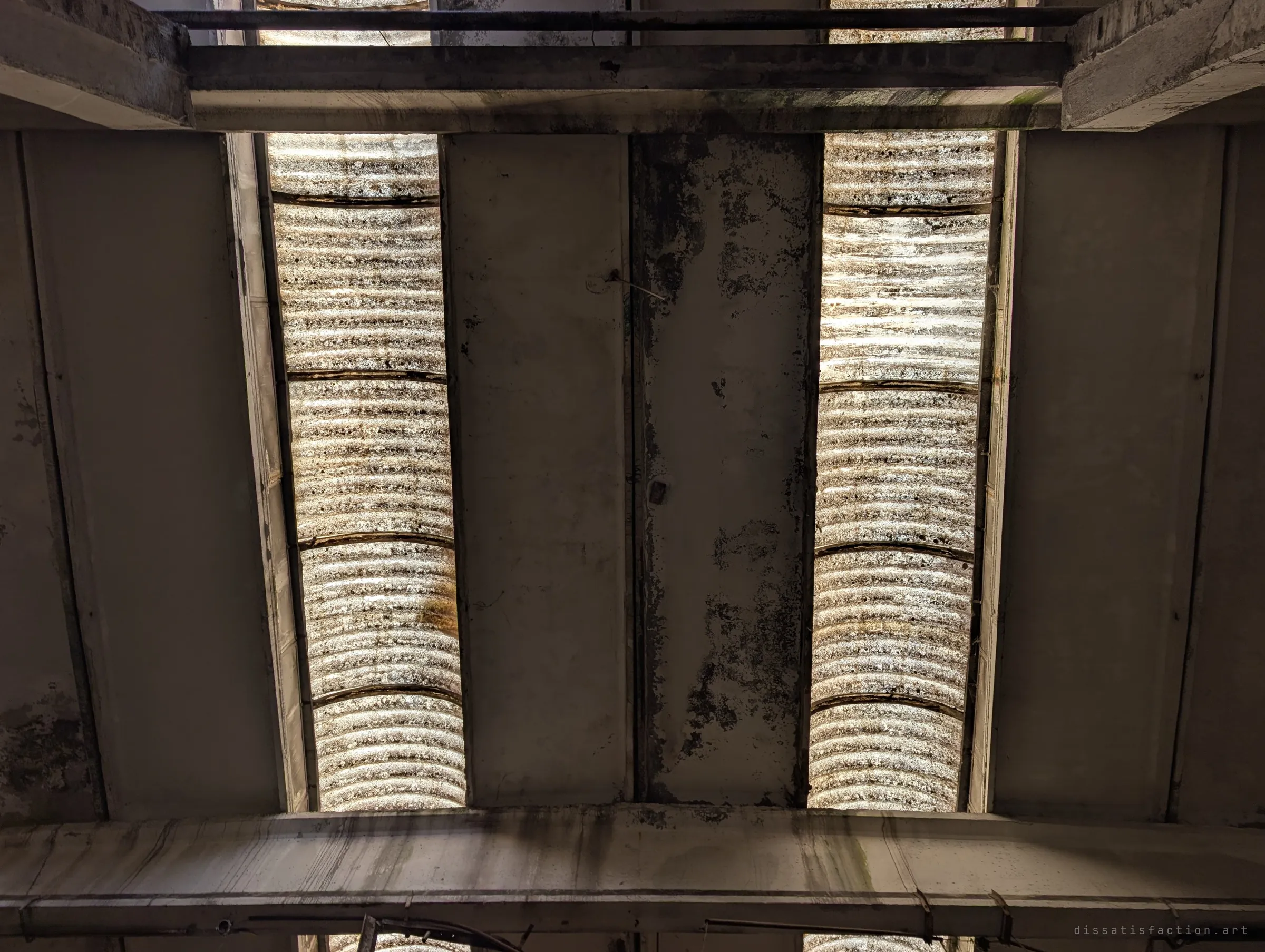 Looking straight up at the roof: two strips of curved, dirt-streaked skylights set between heavy concrete beams. The glow through the grimy panels shows the texture of decades of neglect.