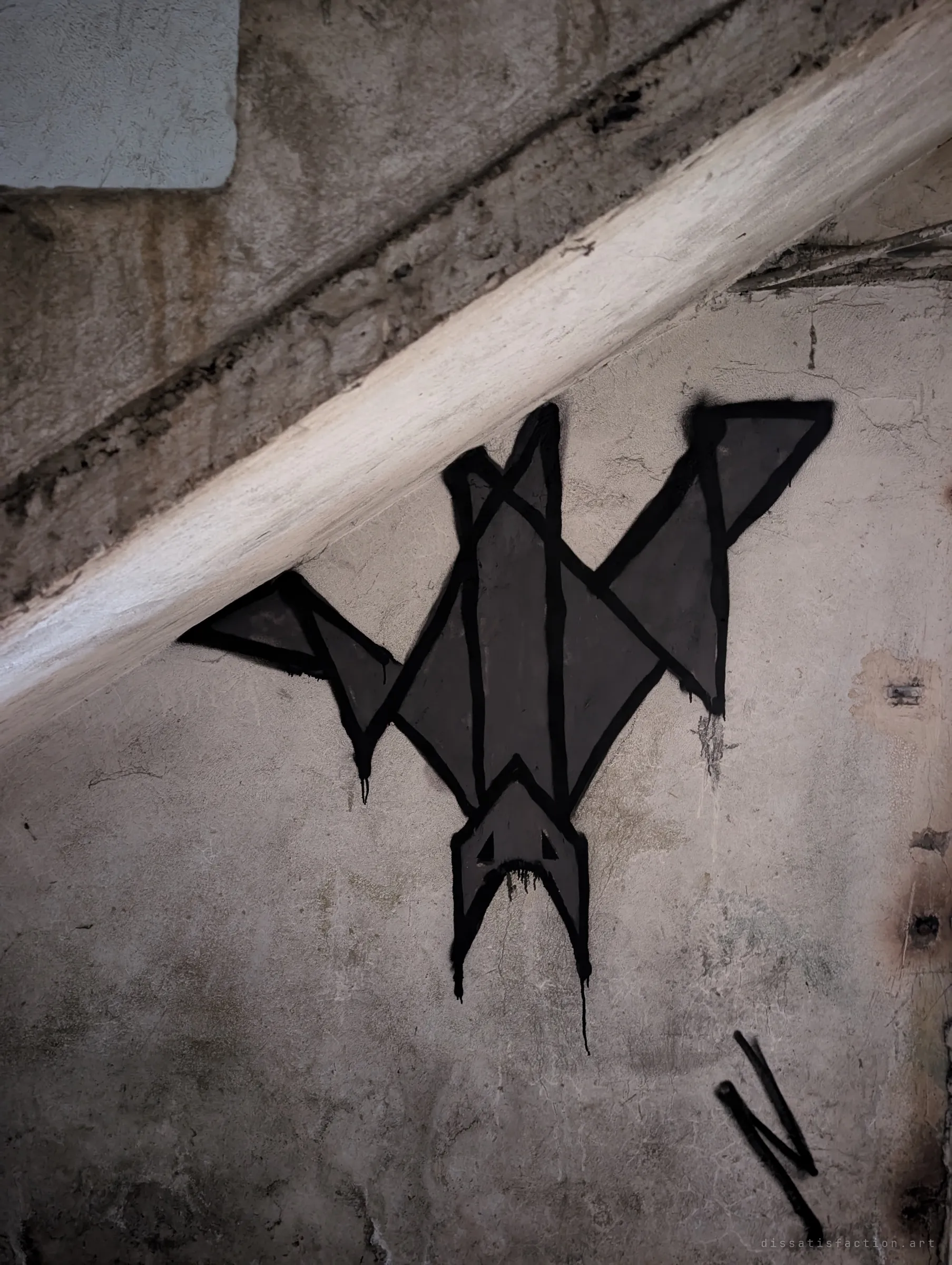 A black geometric bat hangs upside down, spray-painted beneath a diagonal concrete beam. The drips of paint and the stained wall give it an eerie, quiet presence.