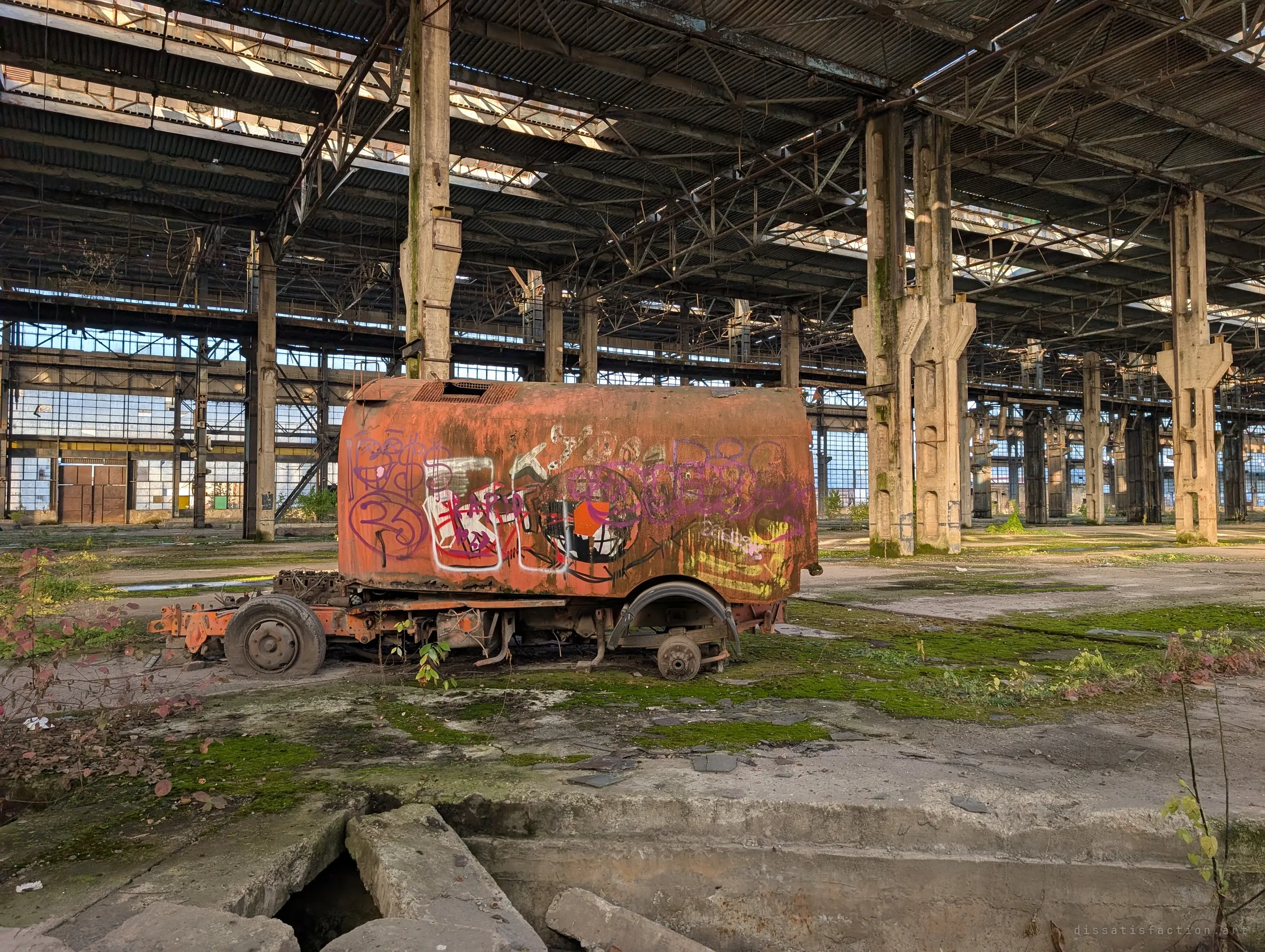 A rusted orange vehicle body covered in graffiti sits on flat tyres in the middle of the hall, framed by tall concrete columns. It is one of the few machines left behind in the vast space.