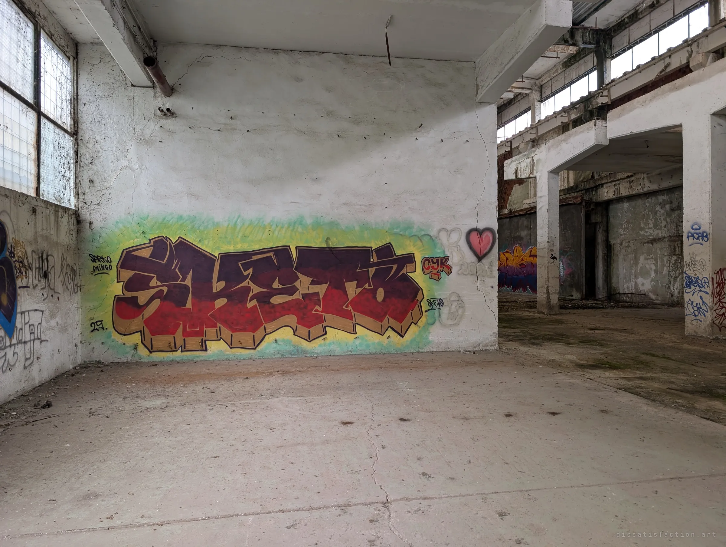 A bold red and brown graffiti piece on a green-yellow cloud fills a whitewashed wall in a corner of the hall, with a small pink heart beside it. To the right, arches lead into further derelict bays.
