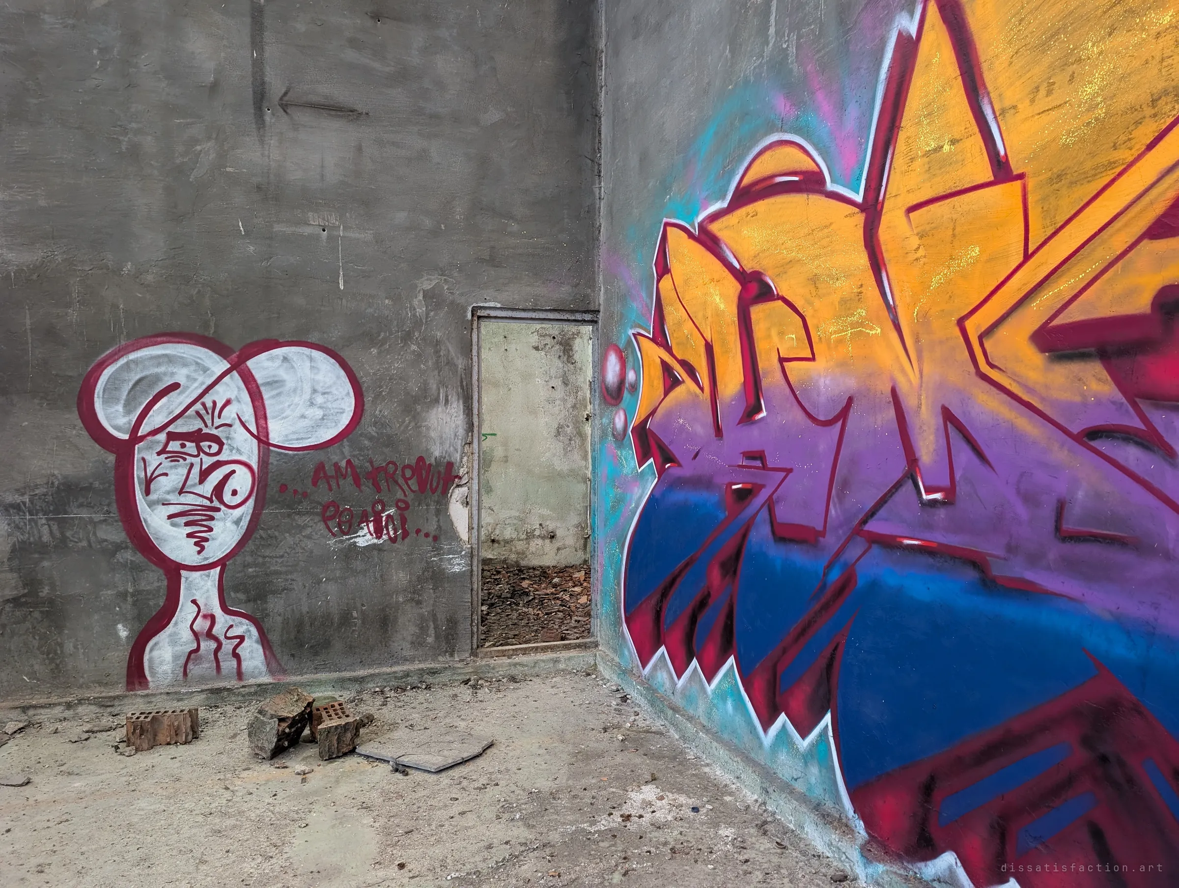A doodled cartoon figure with a short painted phrase stands next to a doorway on a grey wall, facing a vivid orange, purple and blue piece on the adjoining wall. Broken bricks lie on the dusty floor between them.