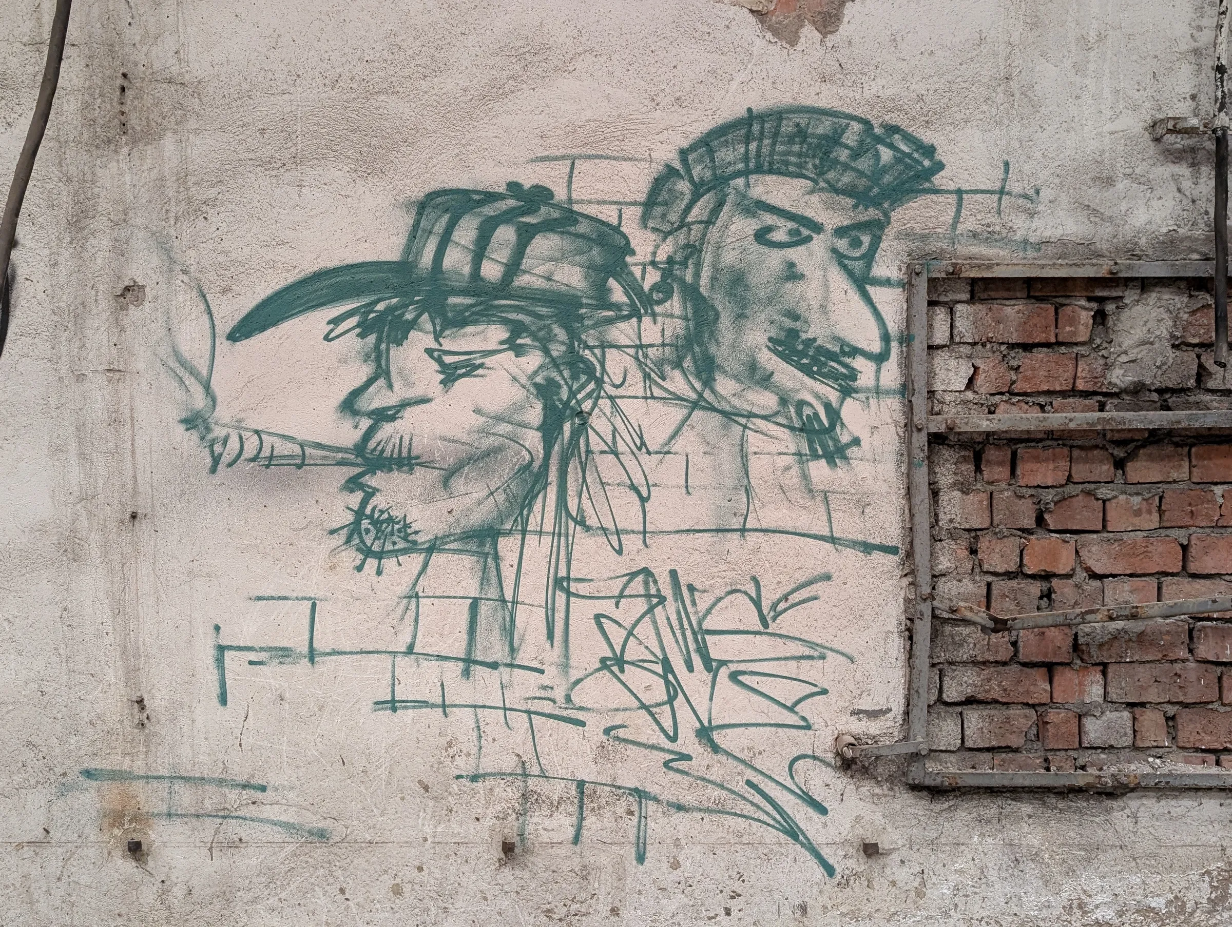 Green spray-paint sketch of two caricatured faces, one with a cigarette, on a peeling plaster wall beside a bricked-up window frame. Casual graffiti that later visitors left on the empty works.
