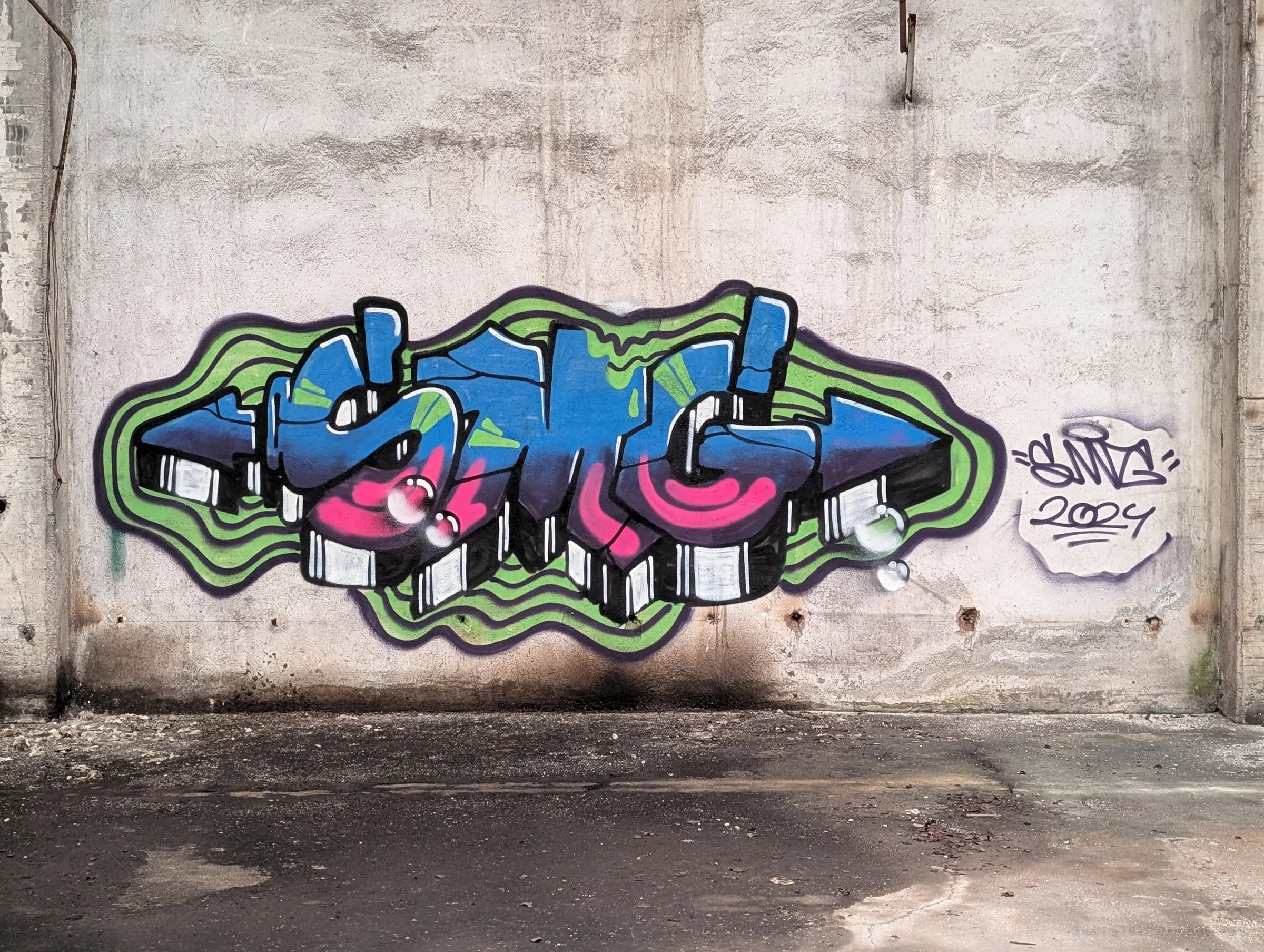 A large blue, pink and green graffiti piece with a dated signature, spread across the bare concrete wall of an empty hall. The abandoned space has become a canvas for writers.