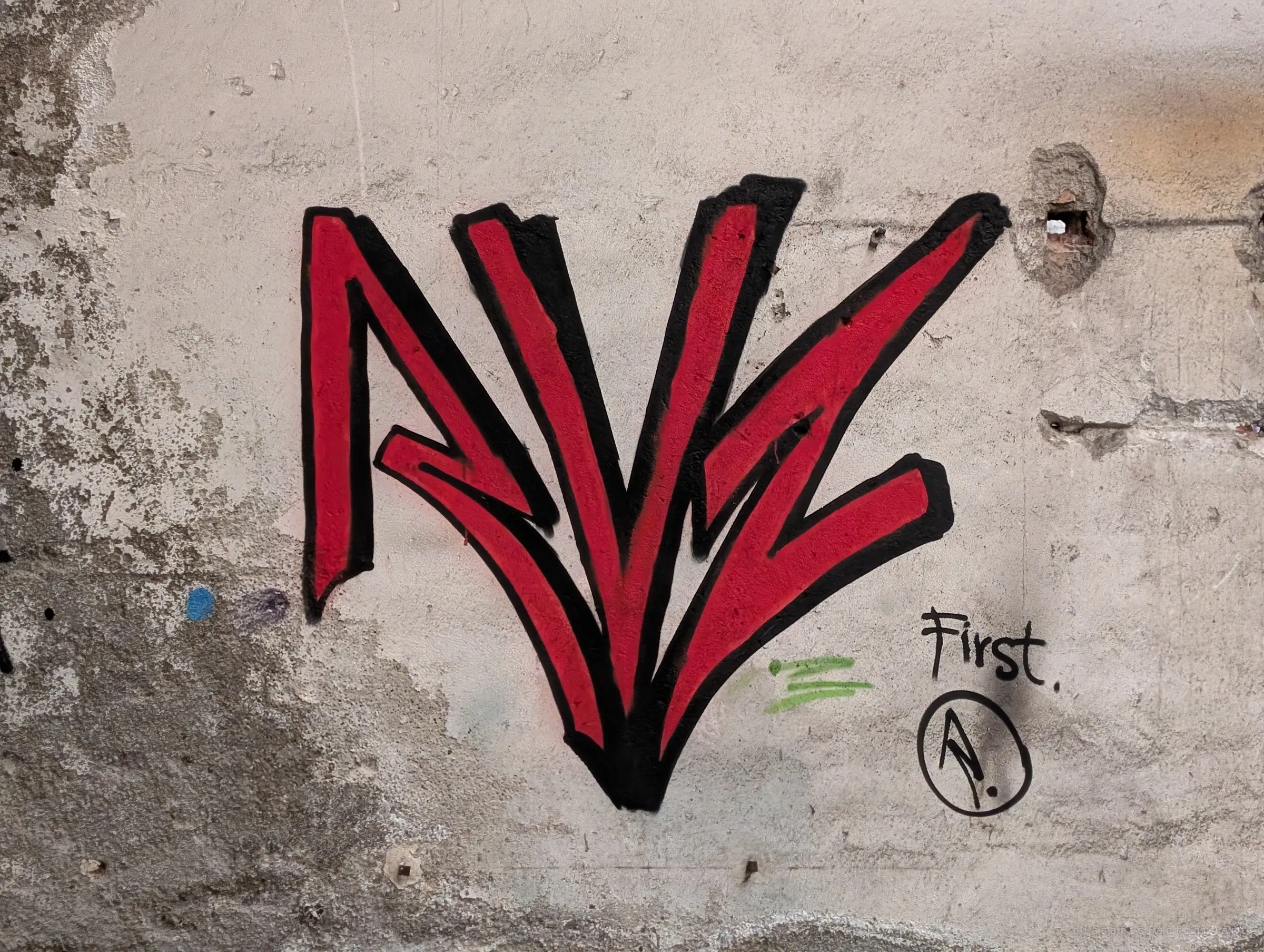 A spiky red-and-black graffiti tag on crumbling plaster, with a small handwritten note and an encircled letter beside it. Detail of the layers of tags covering the ruin.