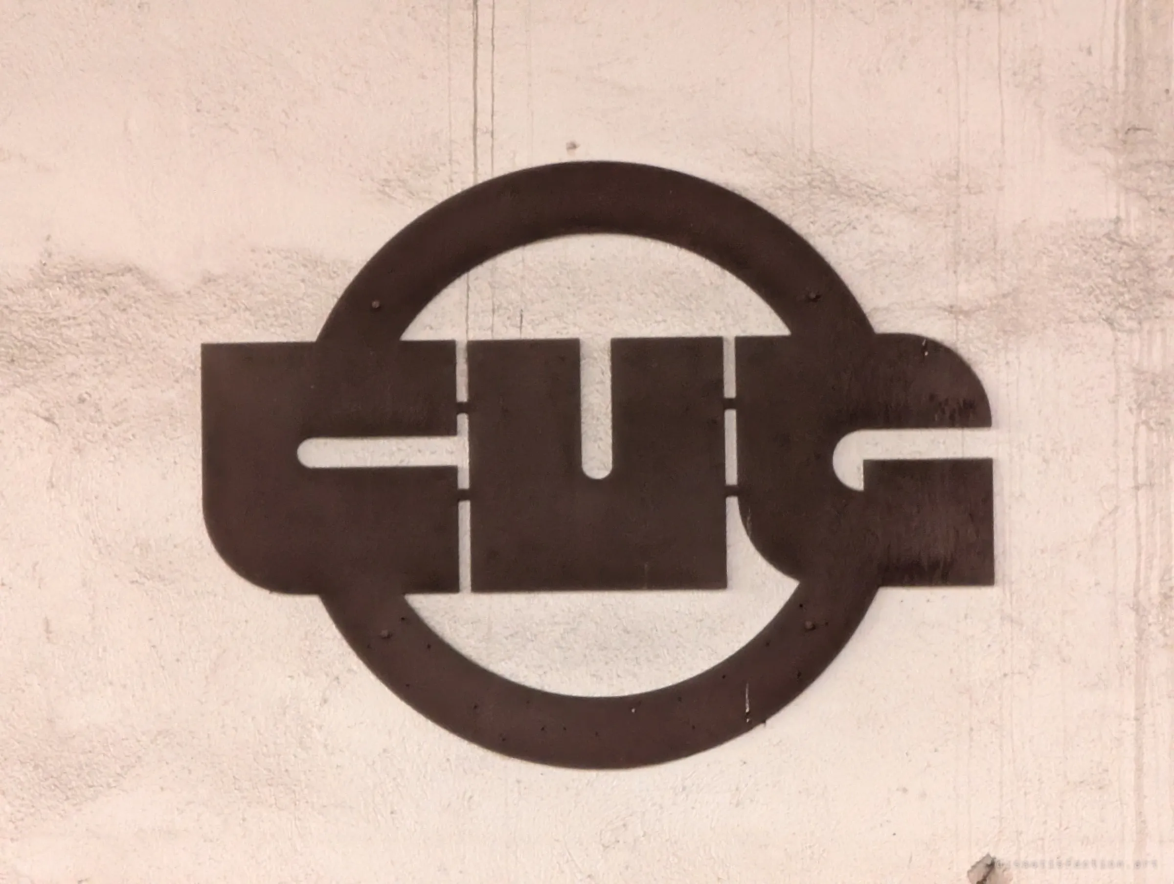 A rusted steel company emblem, block letters inside a ring, still bolted to a pale concrete wall. One of the last marks of the works' original identity.