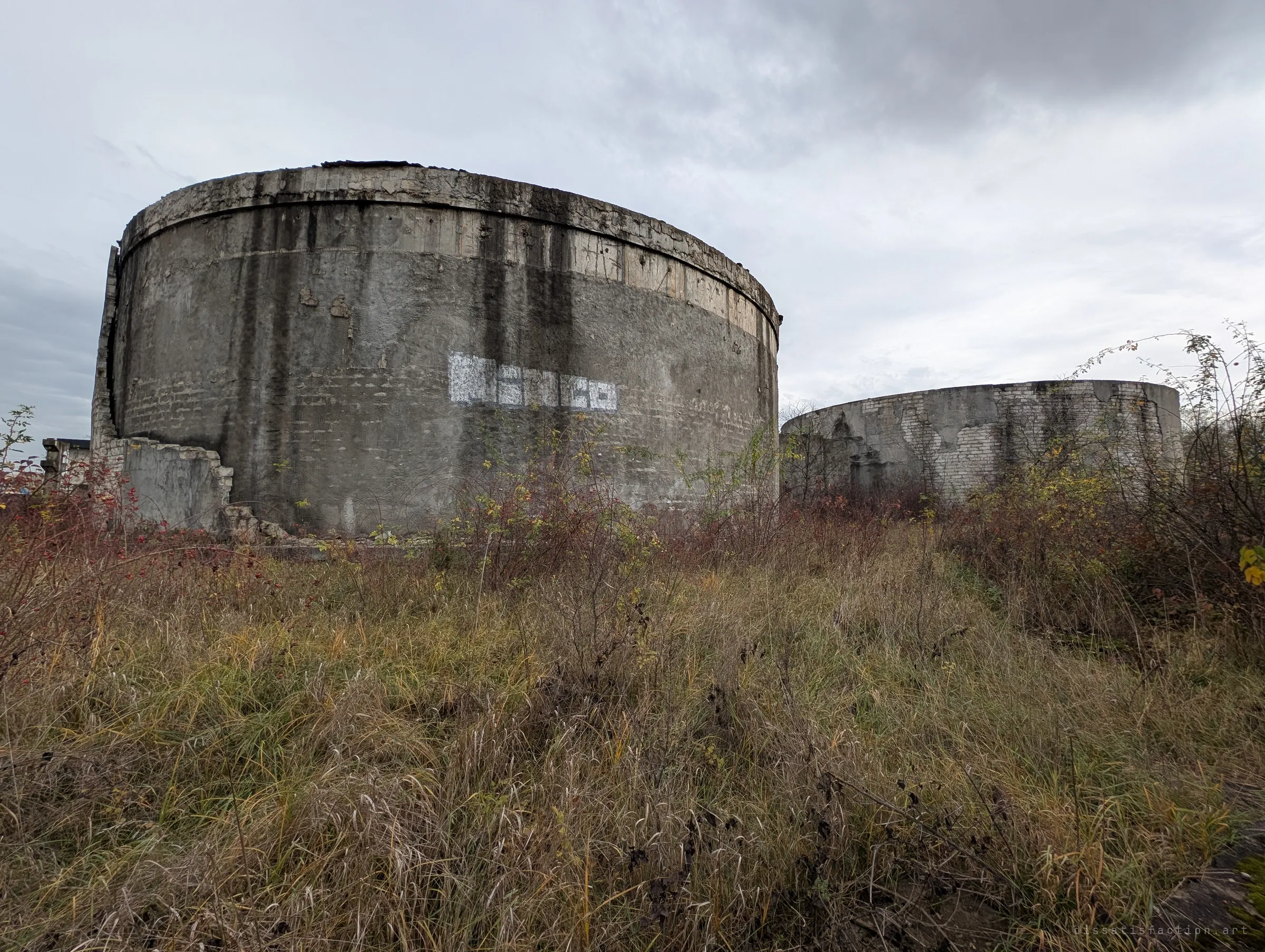 Two roofless round concrete tanks rise from tall autumn grass under a grey sky, one marked with faded white paint. Their streaked walls show decades of weather.