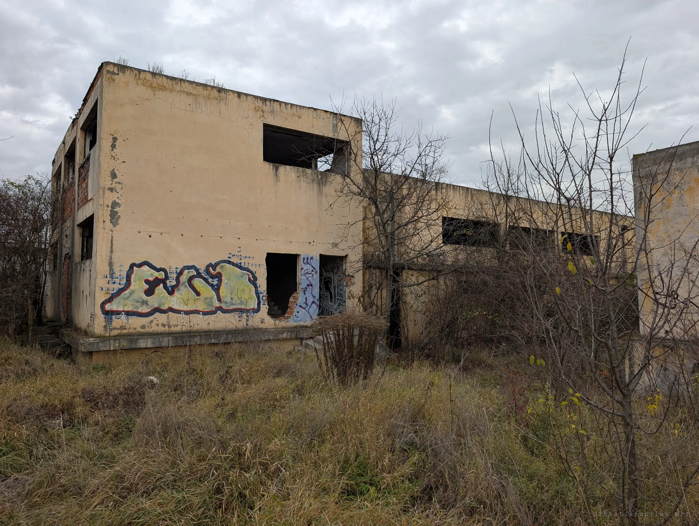 A two-storey yellow-rendered block with empty window openings and graffiti, half hidden behind bare trees and dry grass. A typical outbuilding of the works, stripped to its shell.