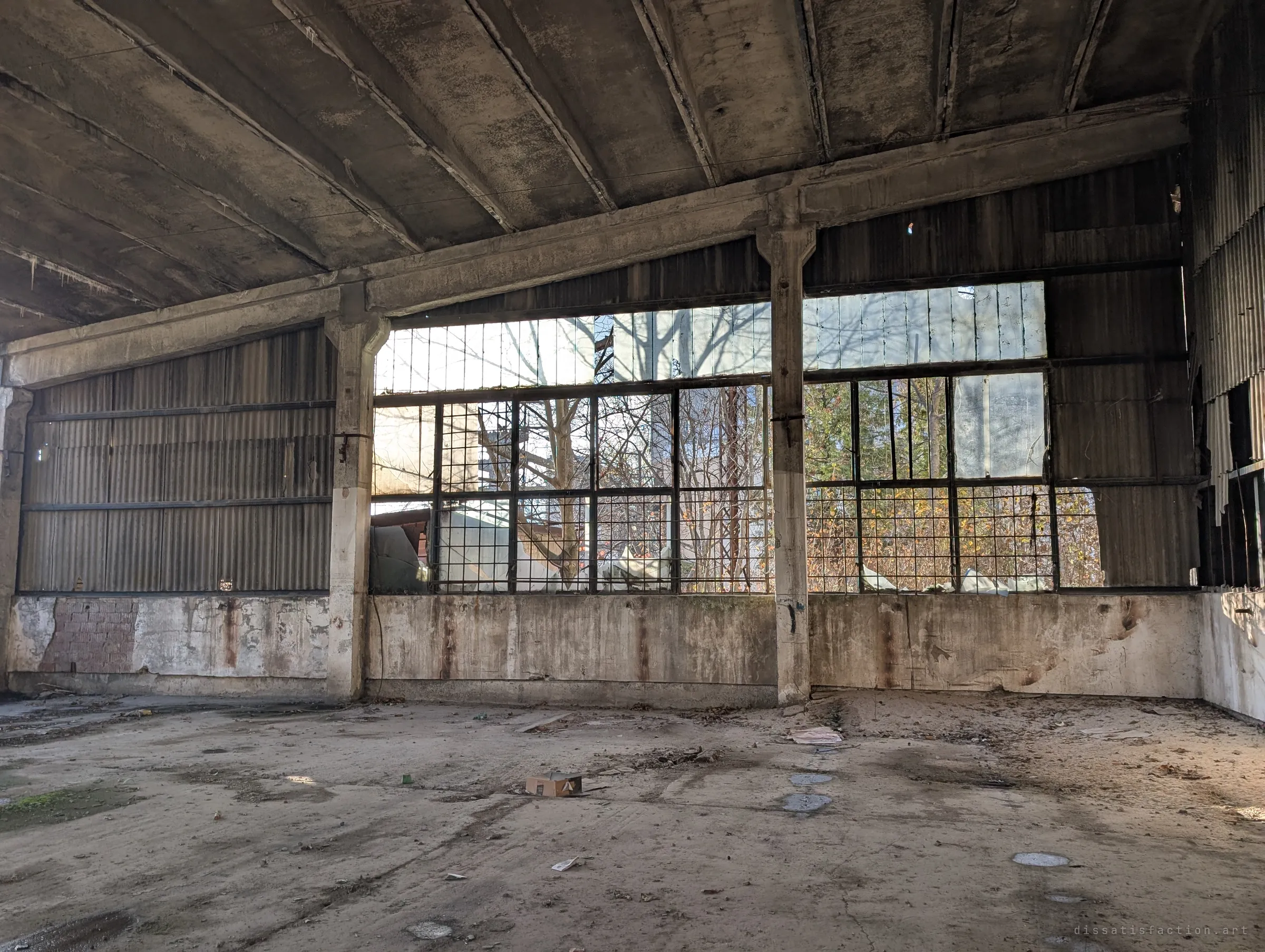 A bare concrete workshop with corrugated walls and a wide barred window looking out onto trees. Low sun lights the dusty floor and scattered paper.