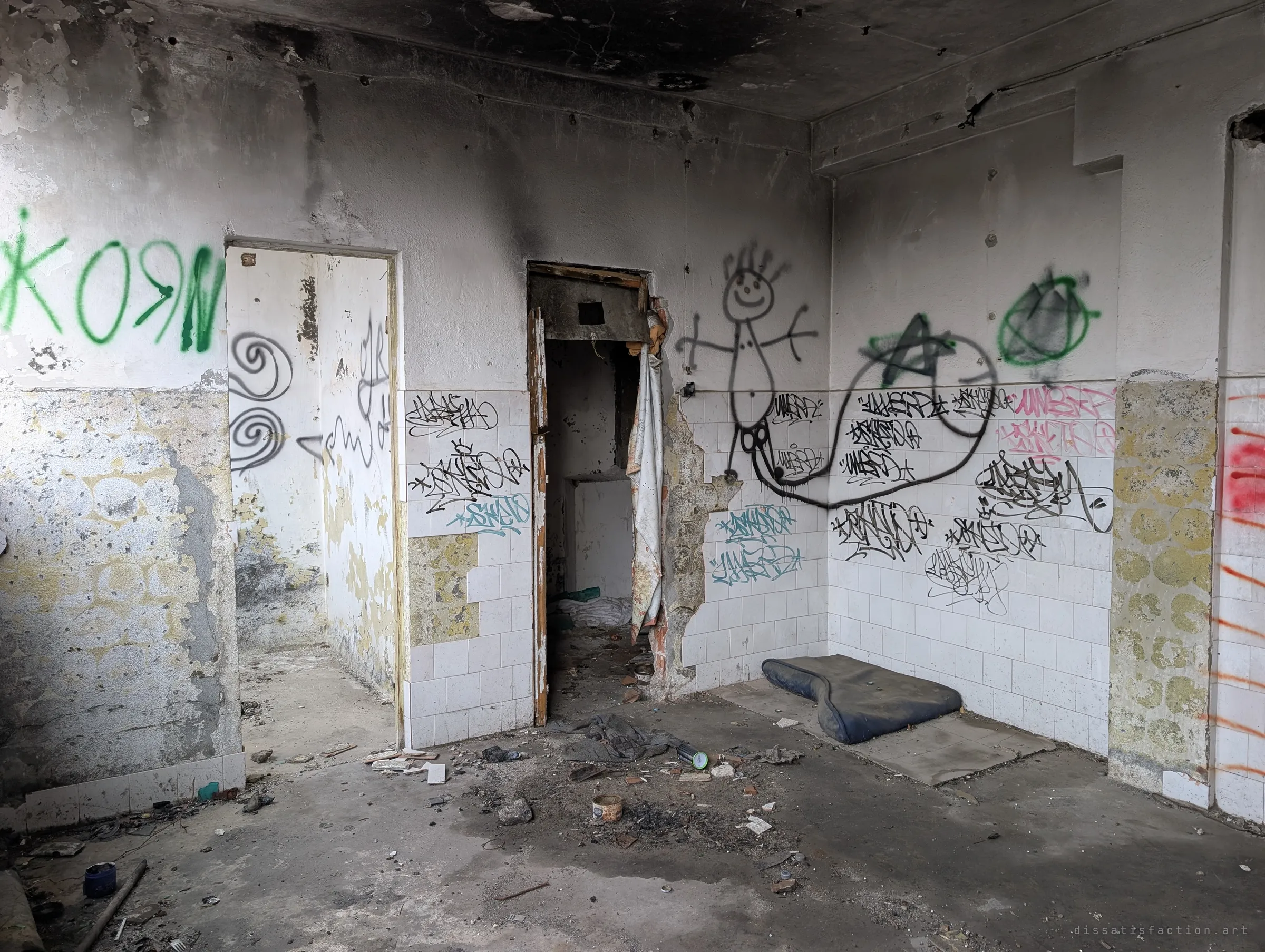 A soot-blackened tiled washroom with tags covering the walls, a stick-figure drawing and a worn mattress pad on the floor. Signs of people sheltering here long after the plant closed.