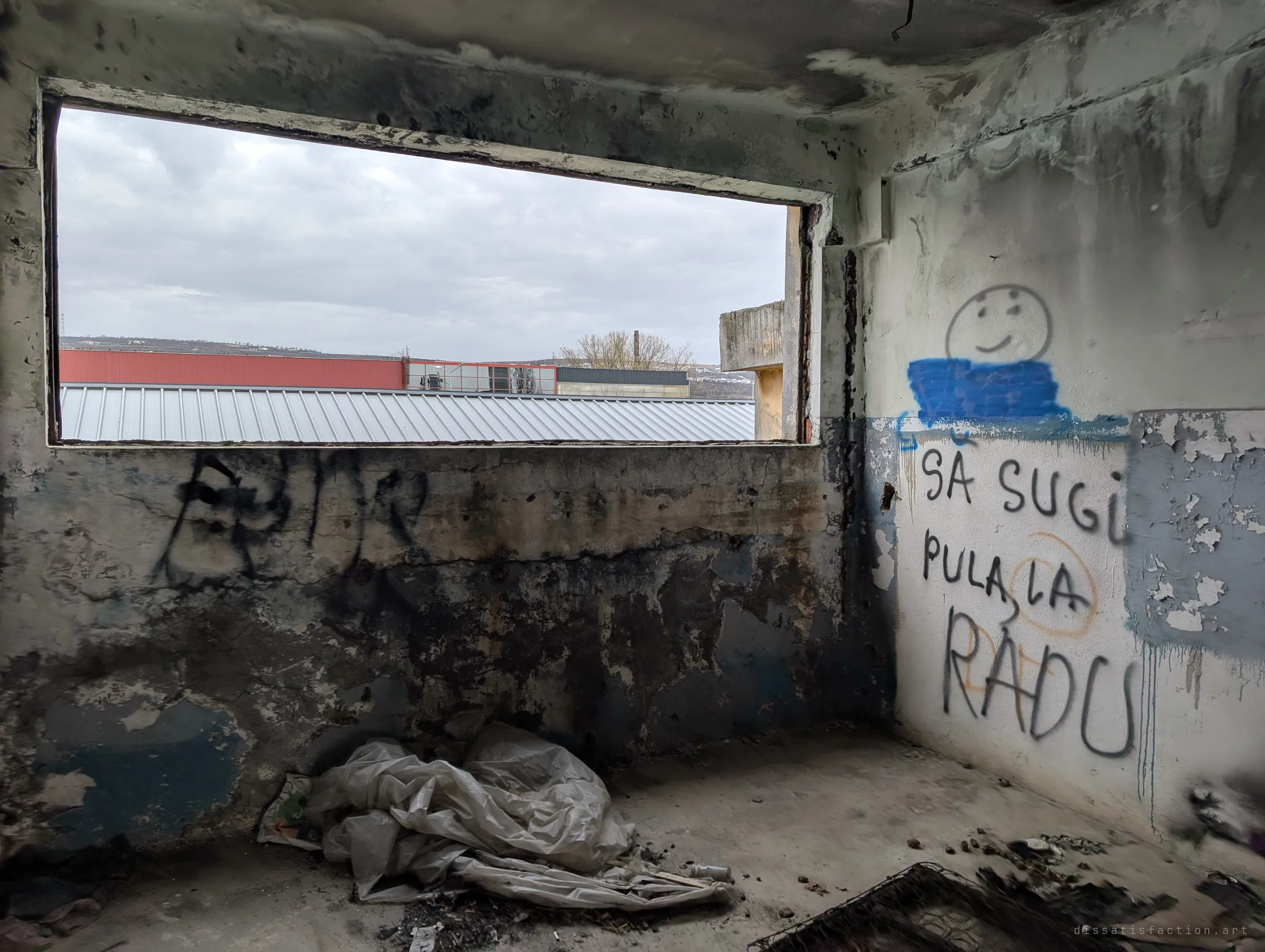 A gutted room with a large empty window framing newer factory roofs and low hills under a grey sky, crude graffiti on the side wall. The working world carries on just outside the ruin.