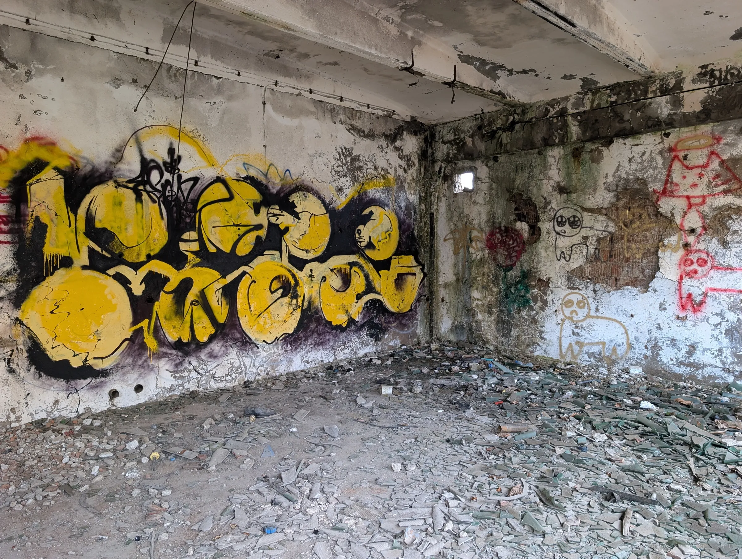 A large yellow and black graffiti piece fills one wall of a bare room whose floor is carpeted with shattered glass; childlike figures are sprayed on the mouldy wall beside it.