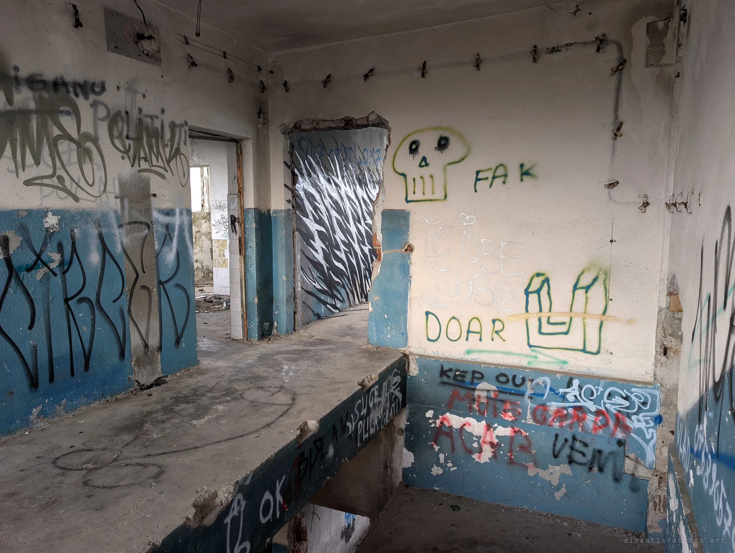 A blue and cream stairwell landing crowded with graffiti: a skull, love notes and a keep-out scrawl, with a doorway opening onto a wall of black and white brushstrokes.