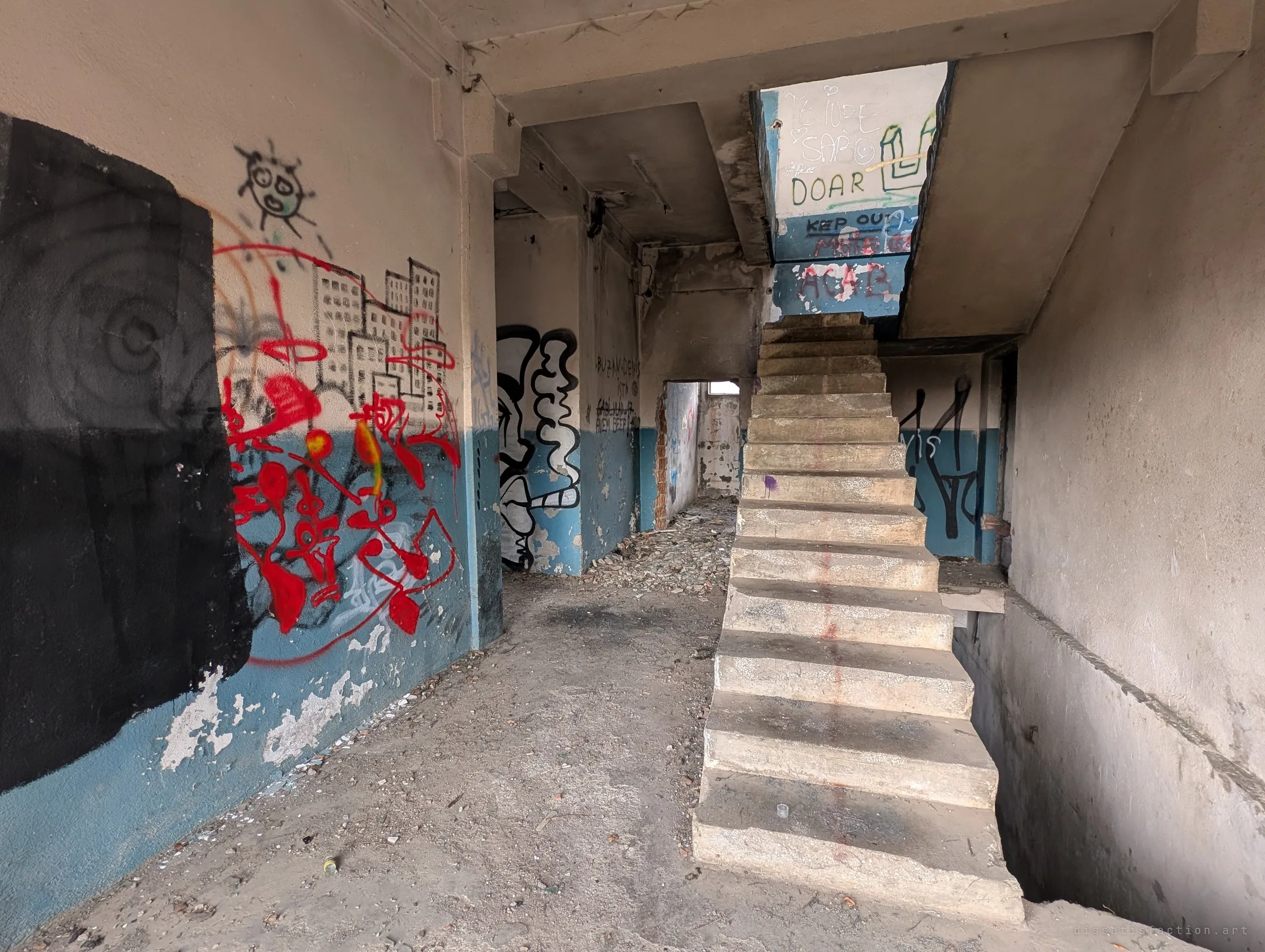 A bare concrete staircase with no railing climbs through a graffiti-covered stairwell toward the landing above, a skyline doodle and red scrawls on the wall beside it.