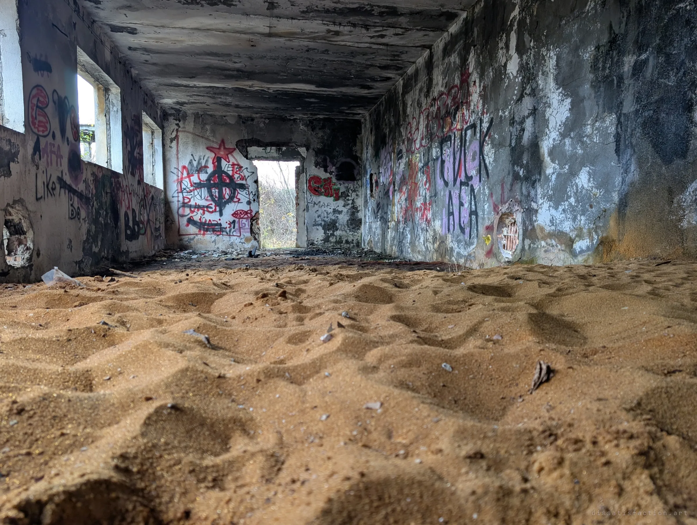 Shot from ground level across a floor buried in rippled orange sand, a graffiti-covered corridor recedes toward a bright doorway. The drifts make the room look like a desert.