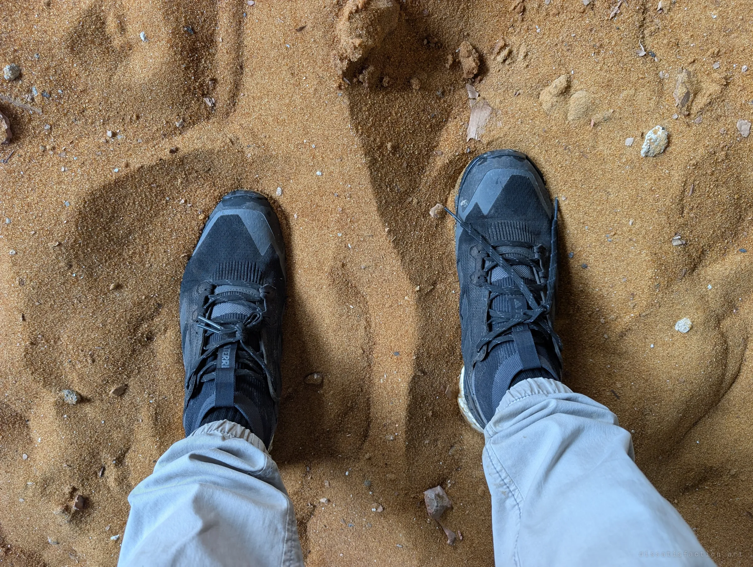 Looking down at black trail shoes sunk into fine orange sand on the building floor, the photographer's own footprints around them.