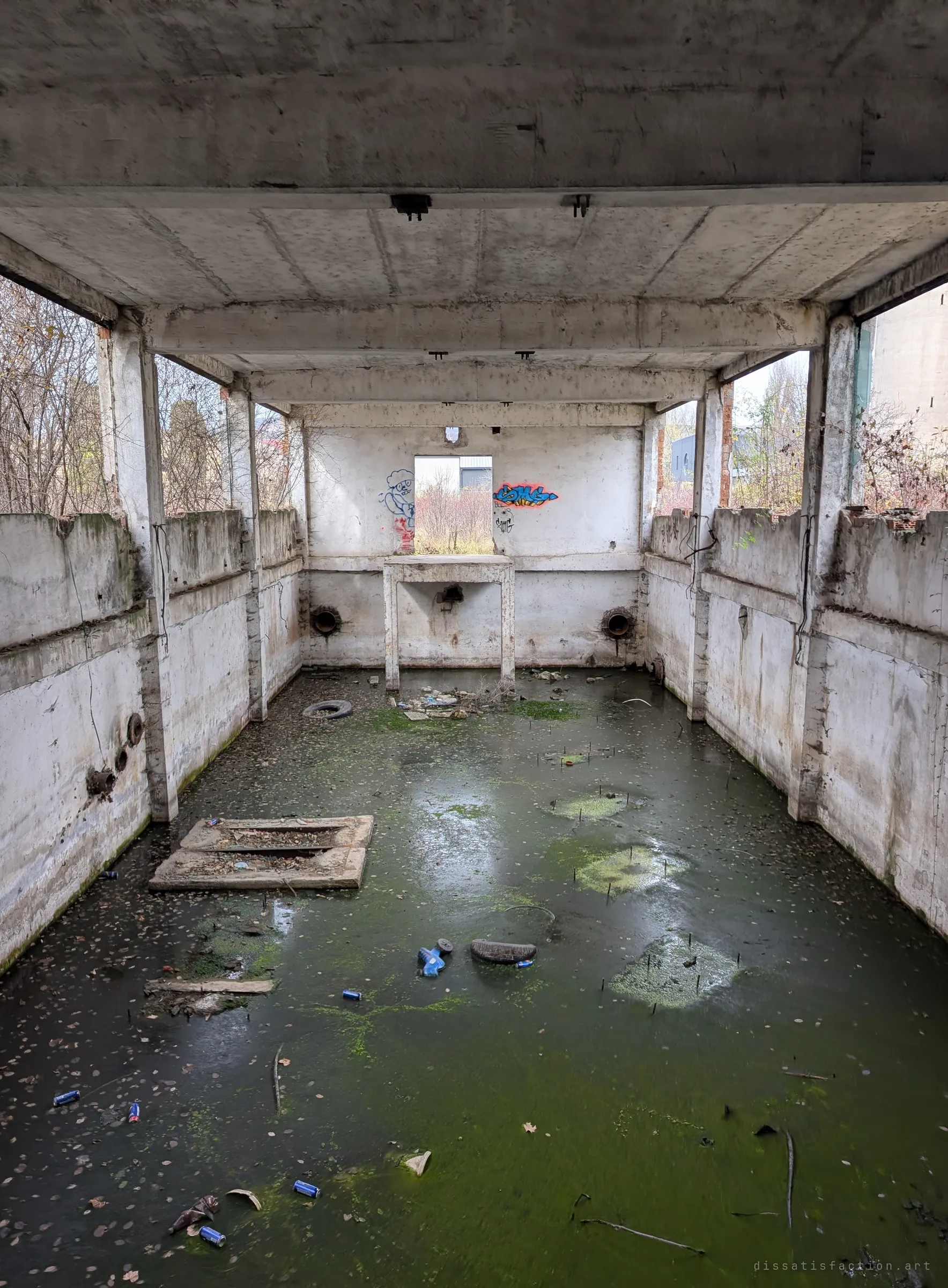 The same flooded concrete basin seen lengthwise in portrait, algae-green water littered with cans and tyres leading to a graffitied back wall with an opening onto overgrown ground.