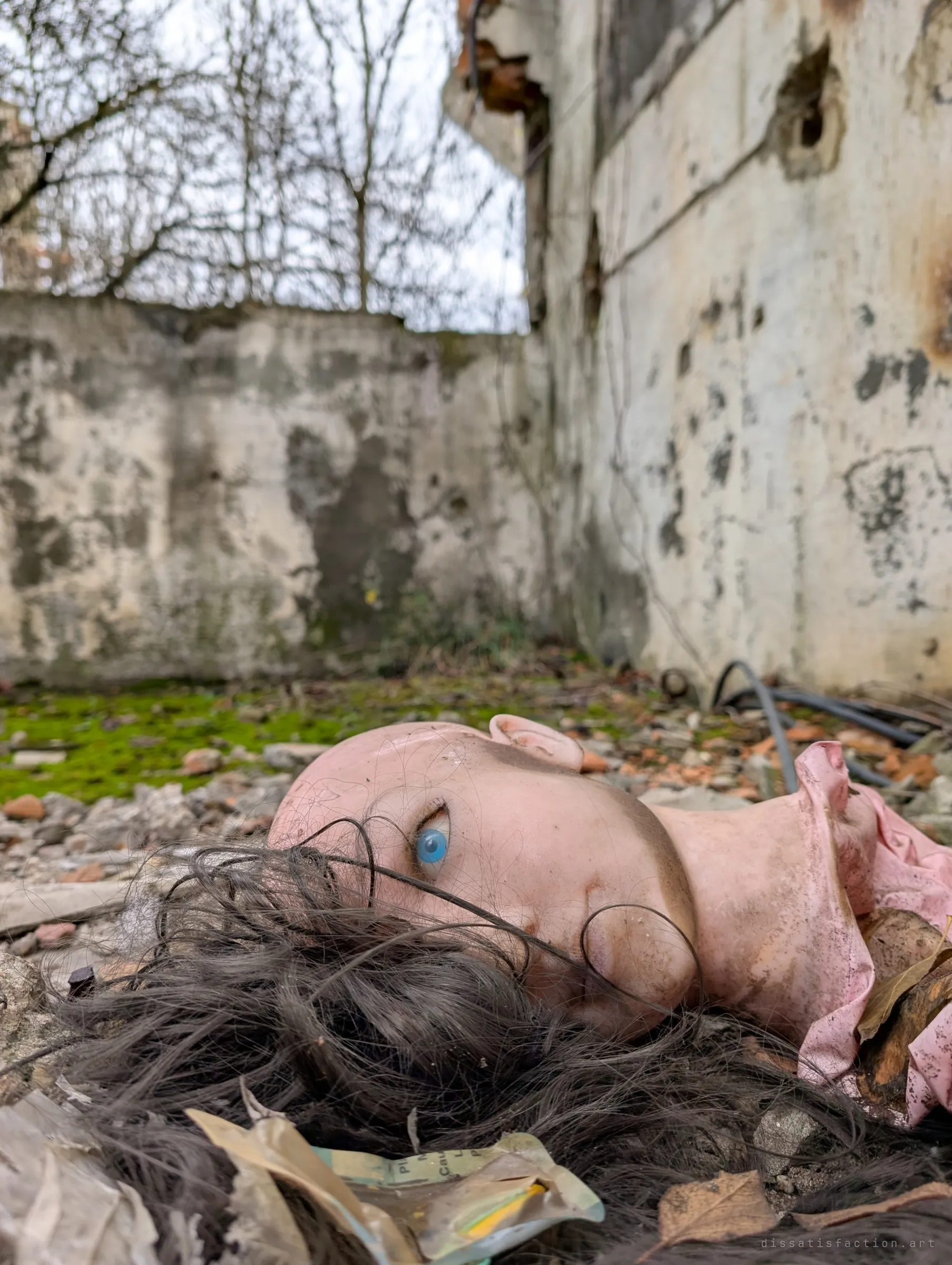 A discarded doll's head with one blue glass eye lies in the rubble, tangled dark hair and a pink collar, with crumbling concrete walls behind. An eerie, human-looking detail among the ruins.