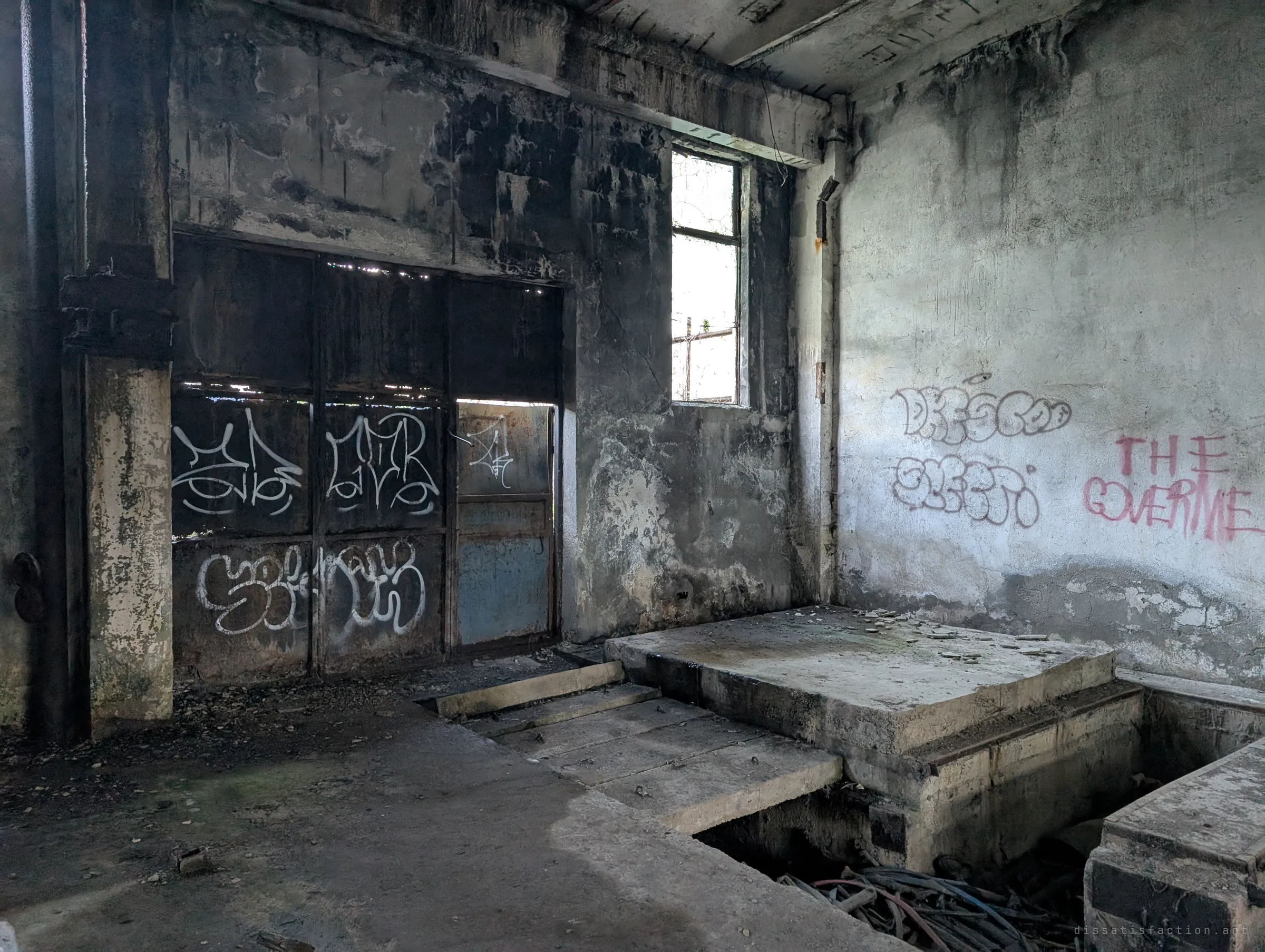 A soot-stained room with a graffiti-tagged steel door, a single window and concrete machine plinths over a pit full of cables. Someone has sprayed a political slogan on the pale wall.