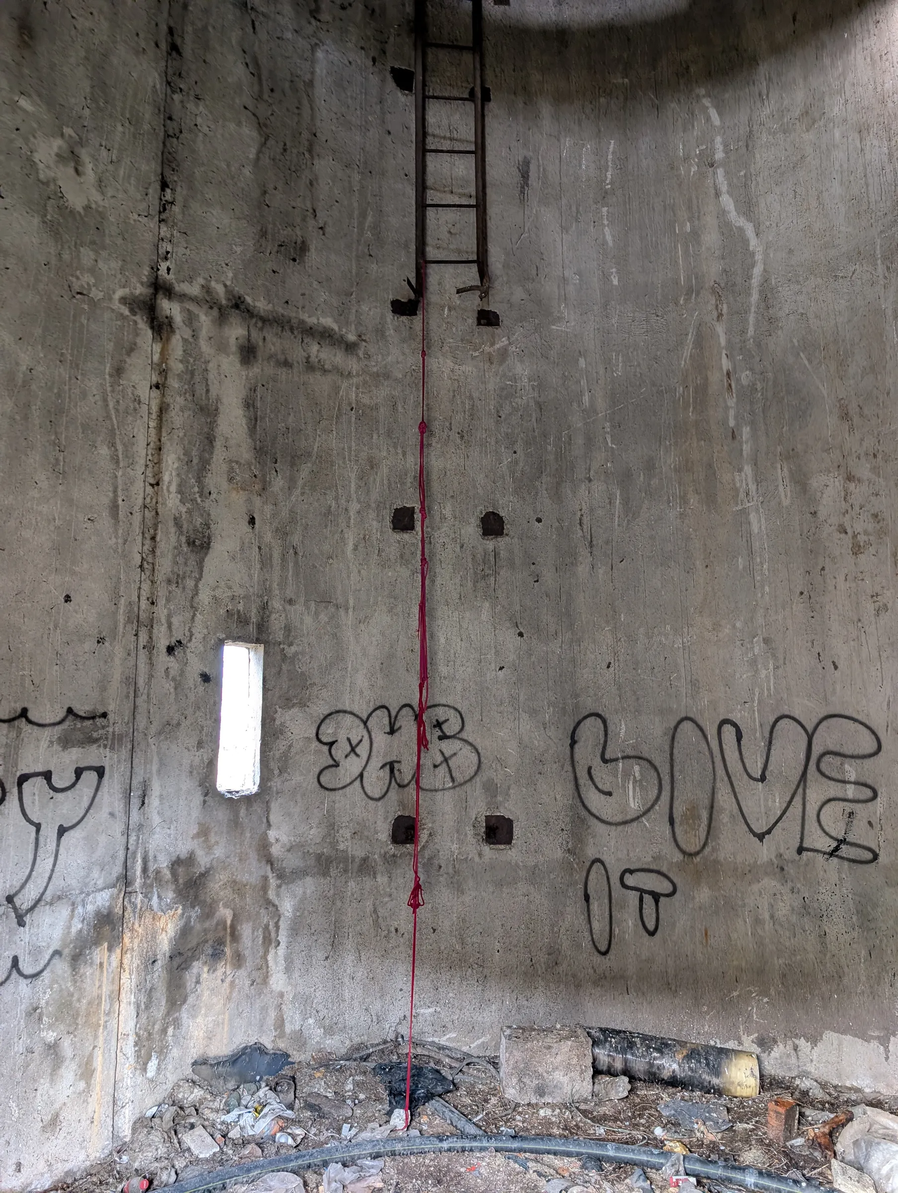 Inside the tower shaft: a rusted ladder begins high on the curved concrete wall, a red rope hanging down from it past graffiti tags and a narrow slit window.