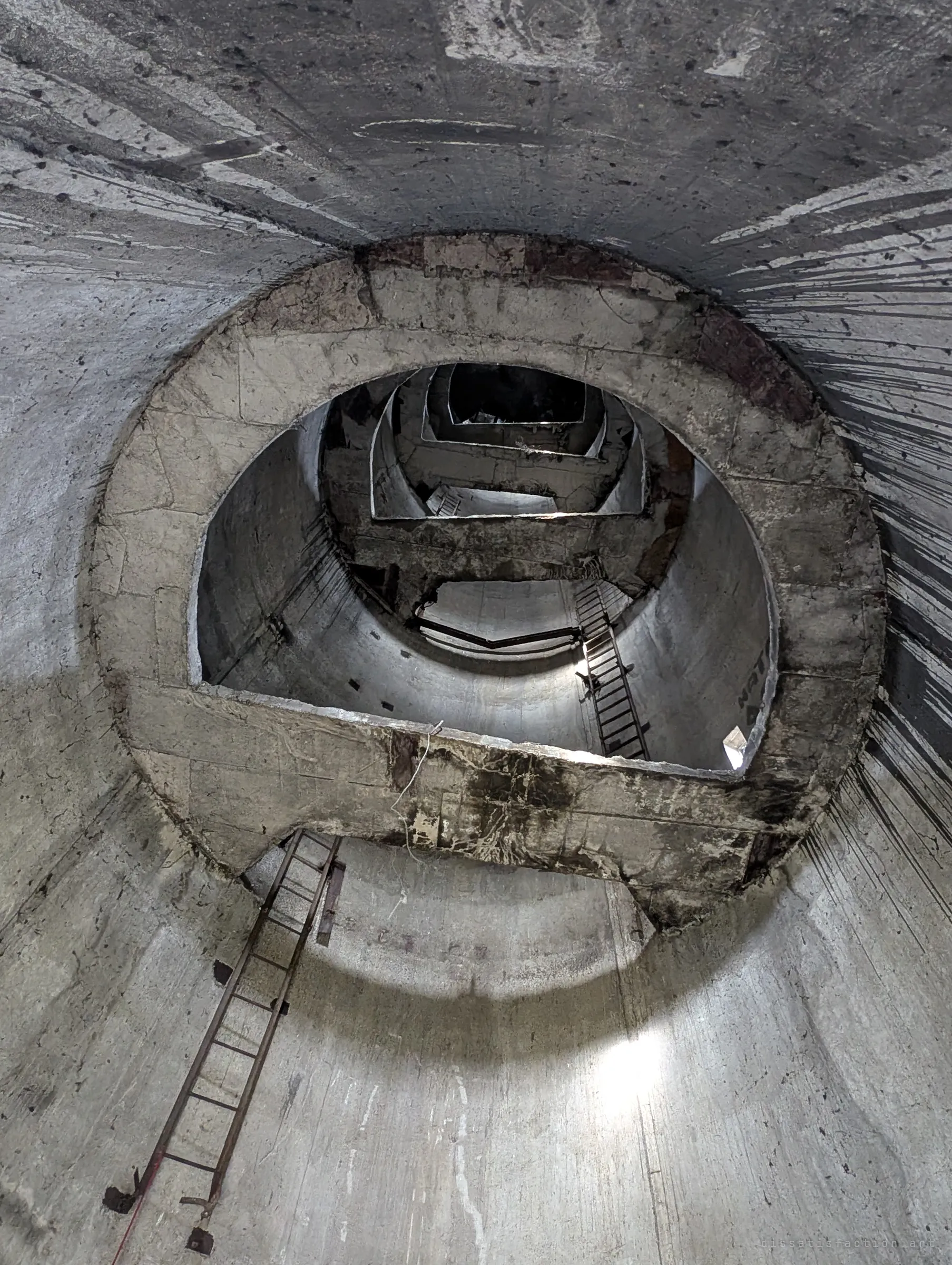 Looking straight up the hollow tower shaft through a ring-shaped concrete platform, ladders climbing toward stacked openings that lead into the tank above.