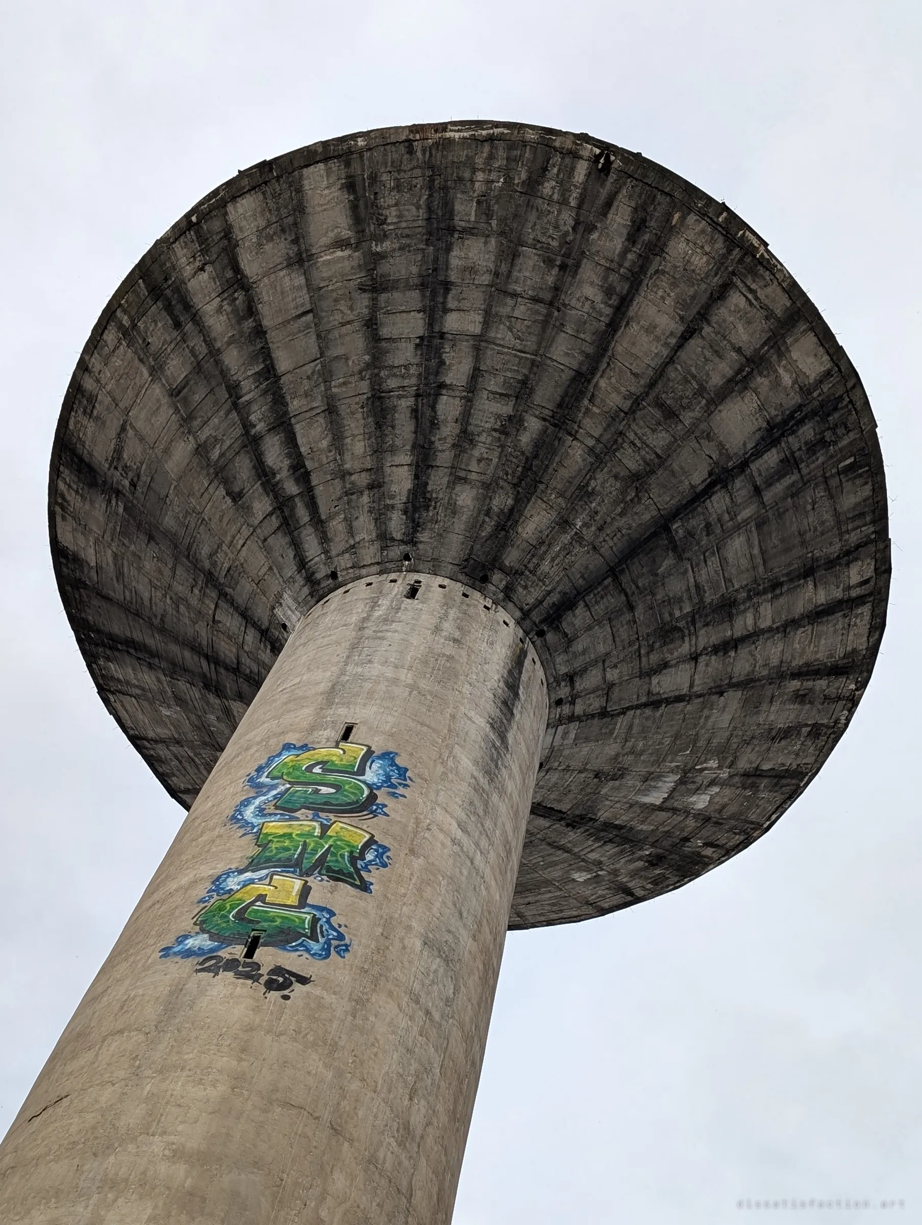 The water tower from directly below, its ribbed concrete tank spreading like a mushroom cap over a shaft painted with a bright green graffiti piece.