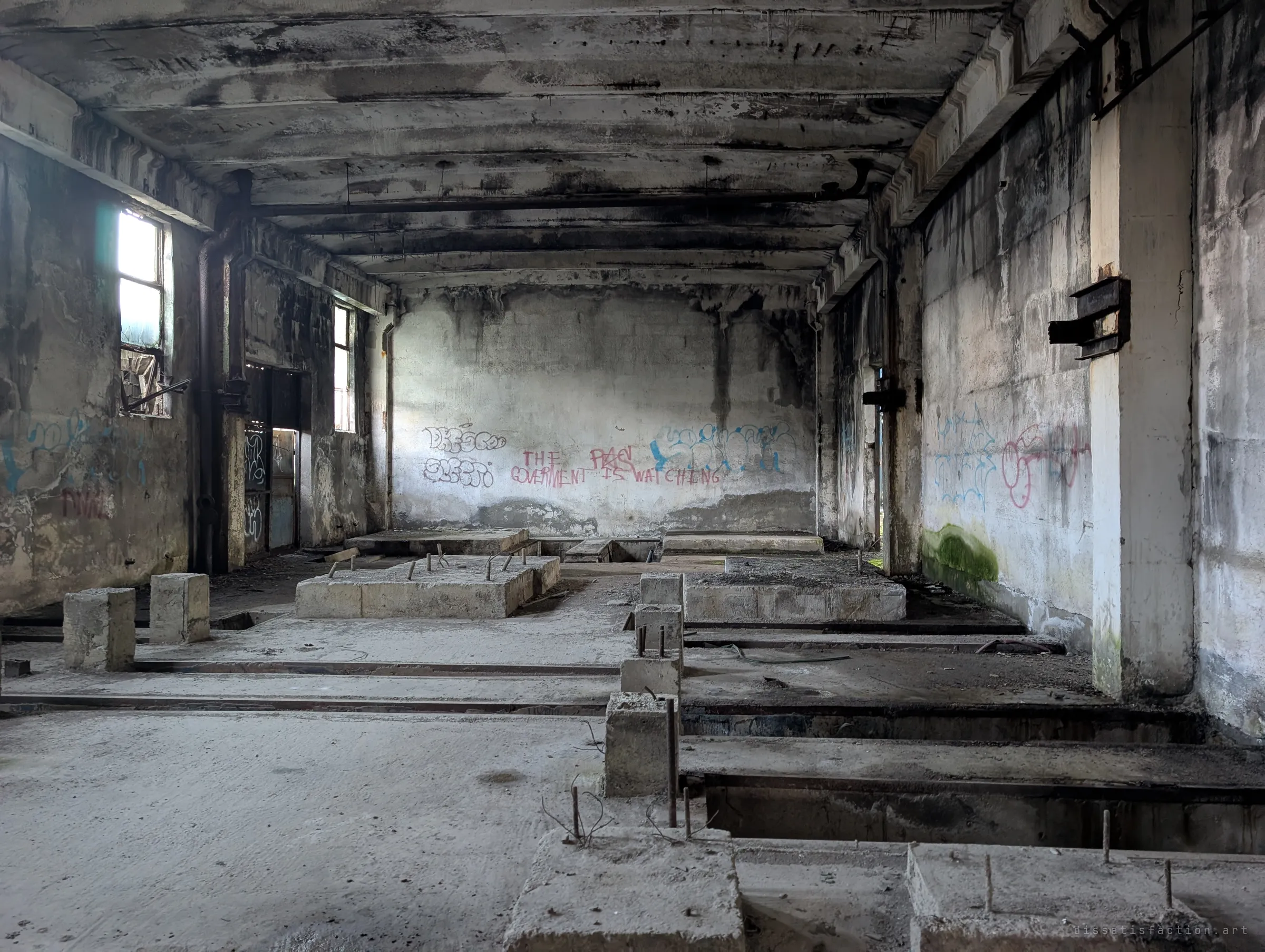 A long blackened hall of empty machine foundations with rebar sticking out of the concrete. Graffiti on the far wall warns that the government is watching.