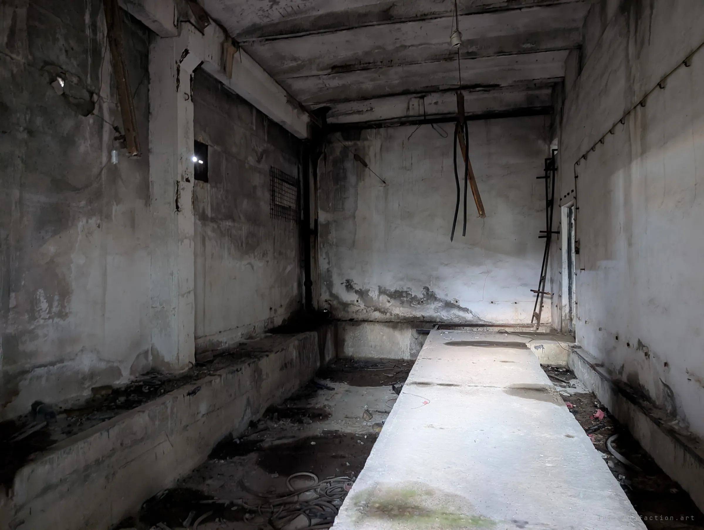A dark concrete room with a raised central slab between two sunken troughs of tangled cable, lit by a flashlight beam. Cut wires hang from the ceiling where equipment was stripped out.