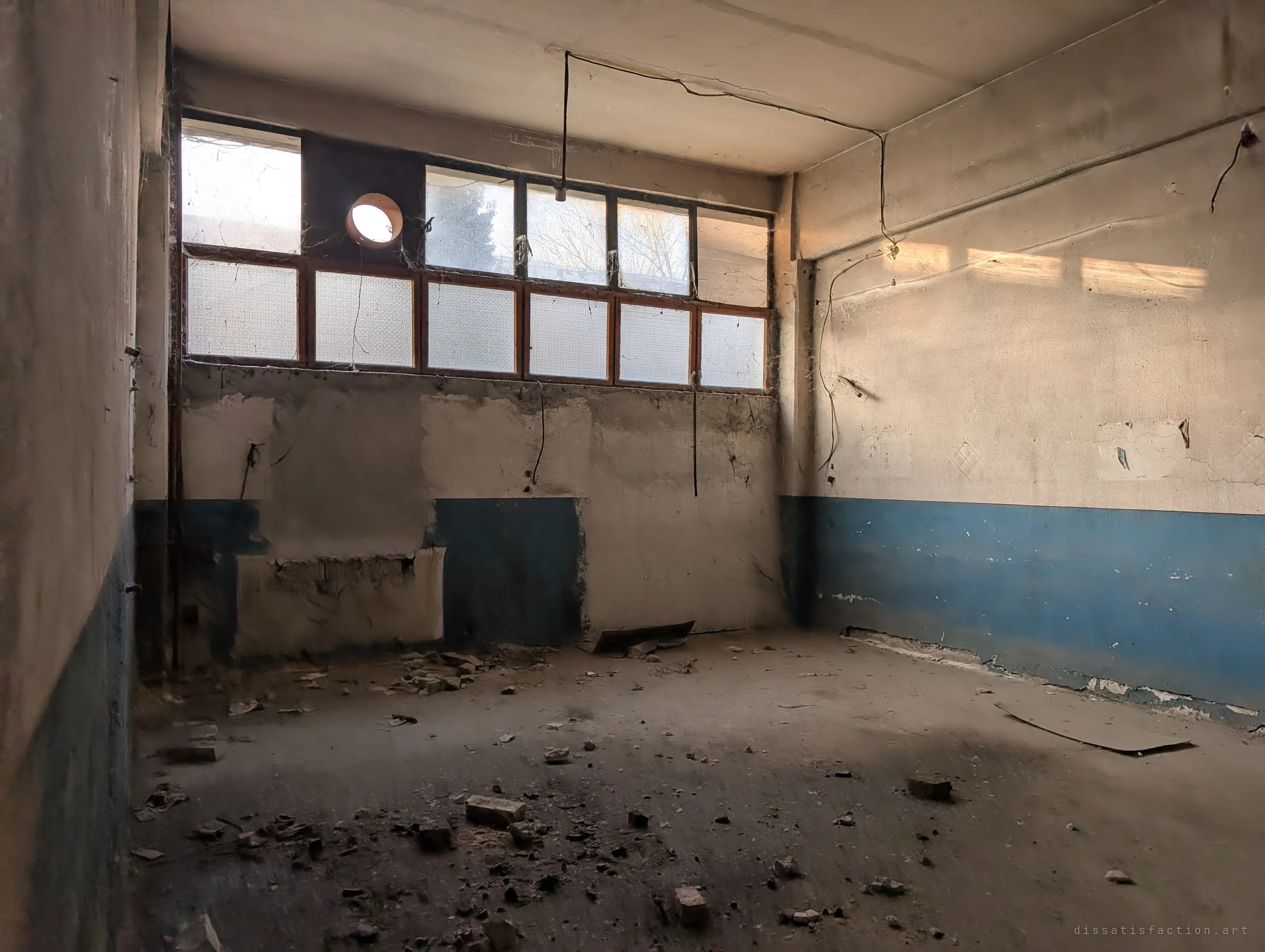 An empty room with a blue-painted dado and a row of frosted clerestory windows, bricks and plaster scattered across the dusty floor. Low sun slants across the wall where cables hang loose from the ceiling.
