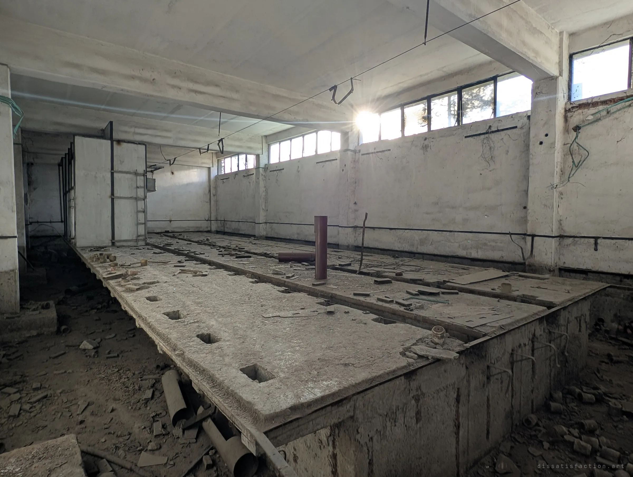 A long workshop hall with a raised concrete bed full of square sockets, littered with metal offcuts, the sun flaring through a high strip of windows. The stripped hall shows where heavy machinery once stood.
