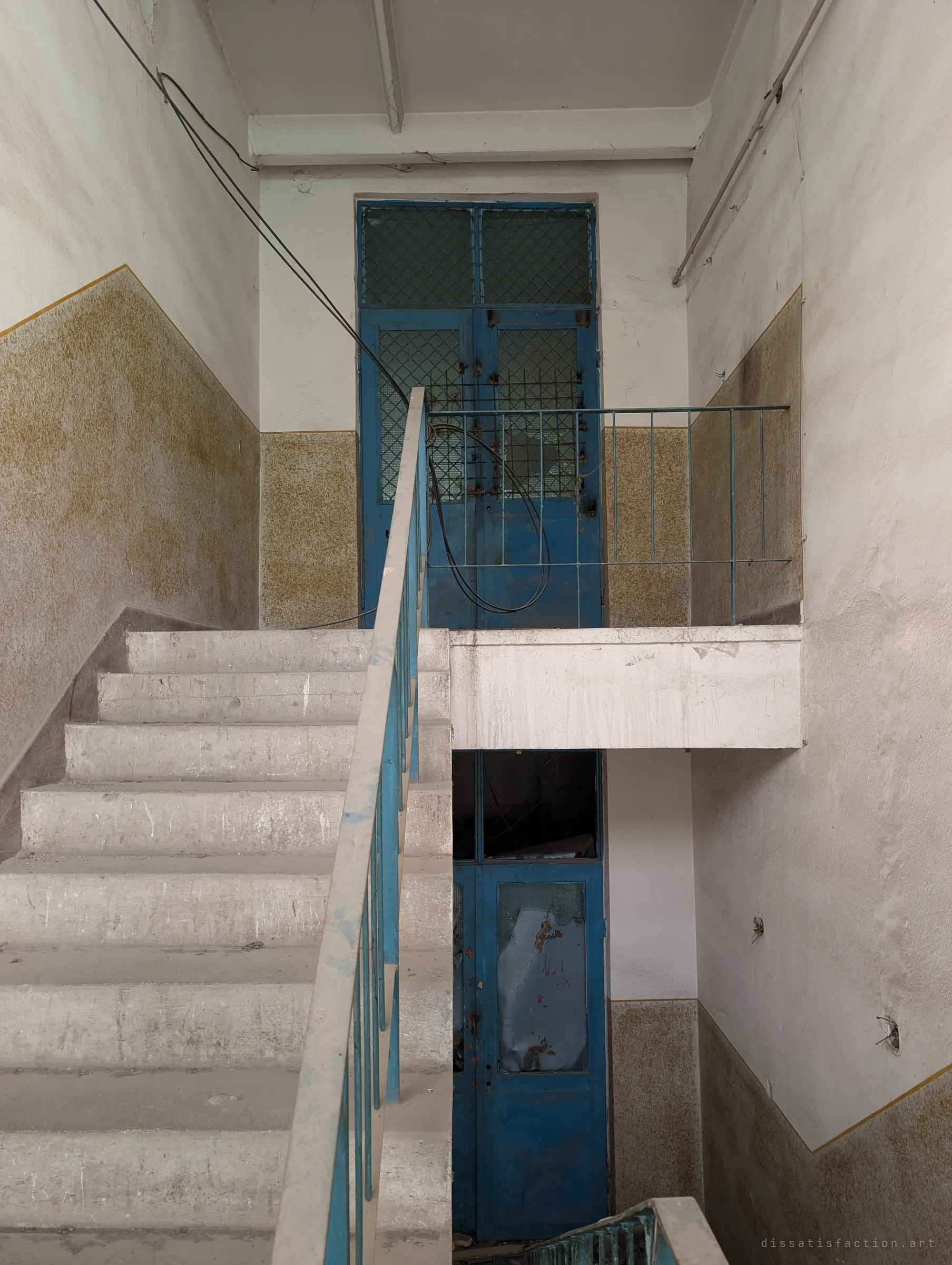 A stairwell with speckled ochre wall paint and blue-painted steel doors with wired glass, a loose cable looping over the railing. The tidy period colours are still intact under the dust.