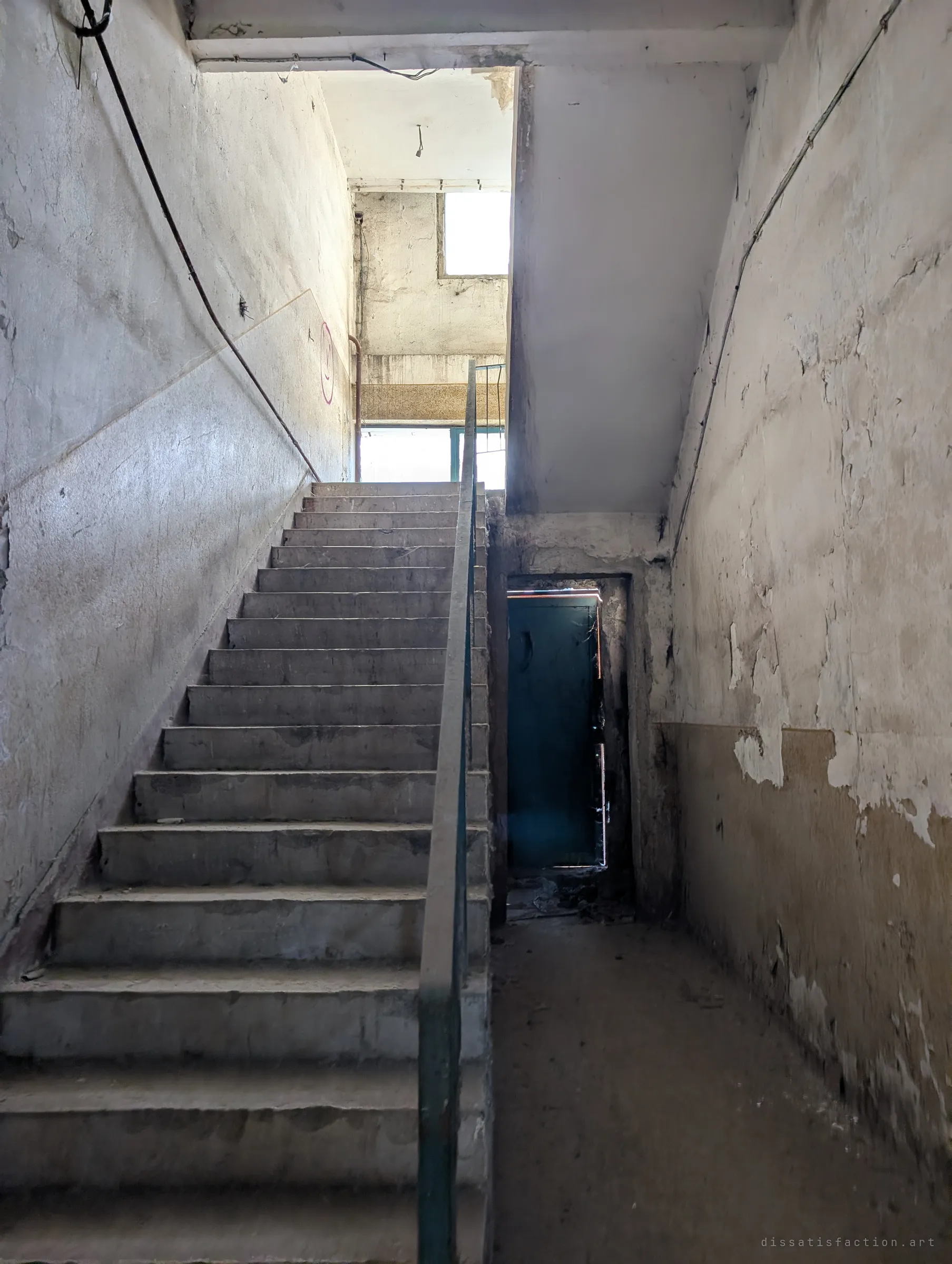 A long concrete stair climbs toward daylight while a dark green door stands half open beside it, light leaking round its edges. Peeling plaster lines the walls of the dim stairwell.