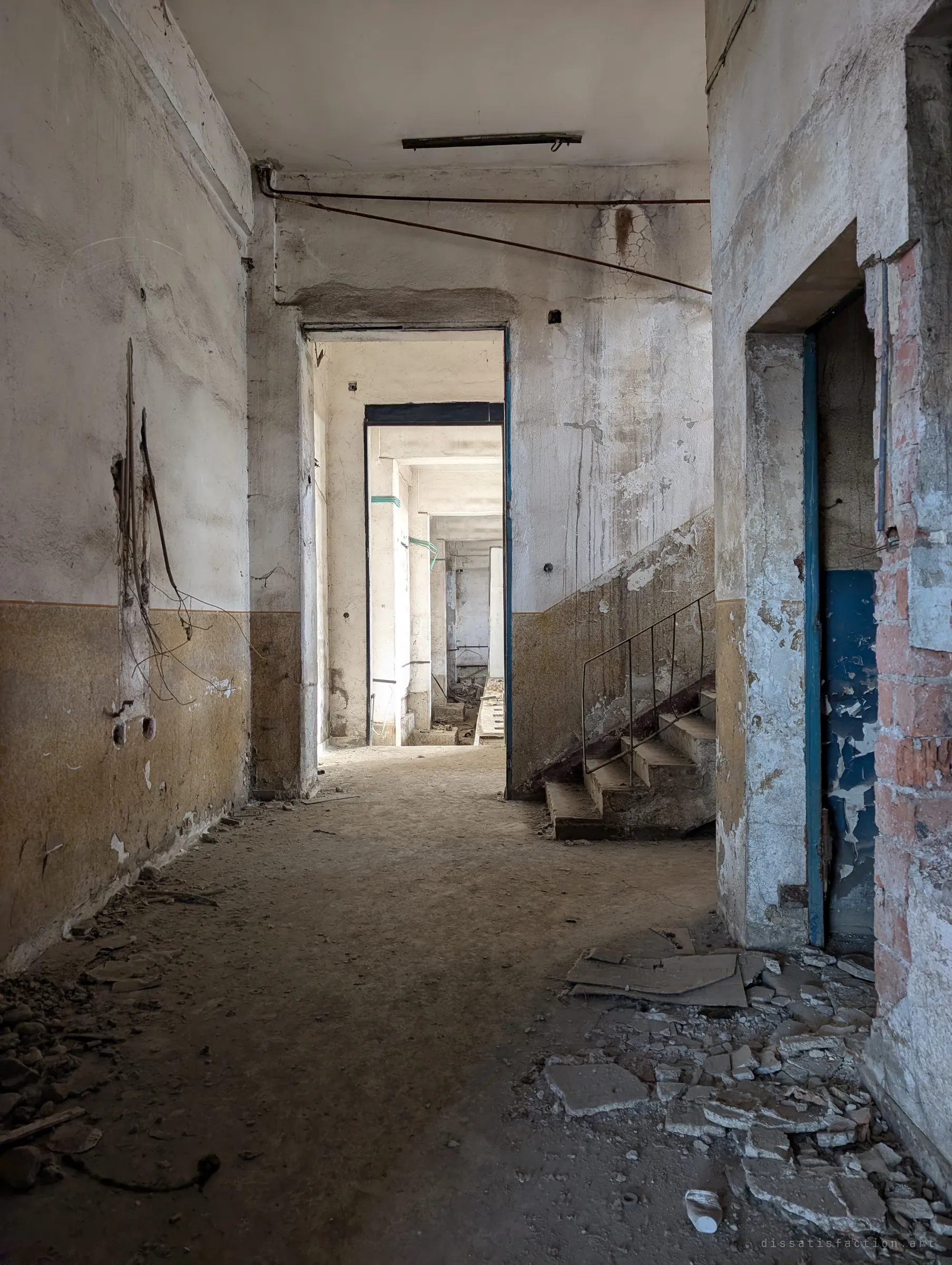 A corridor of cracked plaster and ochre paint leads through a doorway into a chain of bright, empty rooms, a short stair with a thin railing to one side. Rubble and broken tiles cover the floor in the foreground.